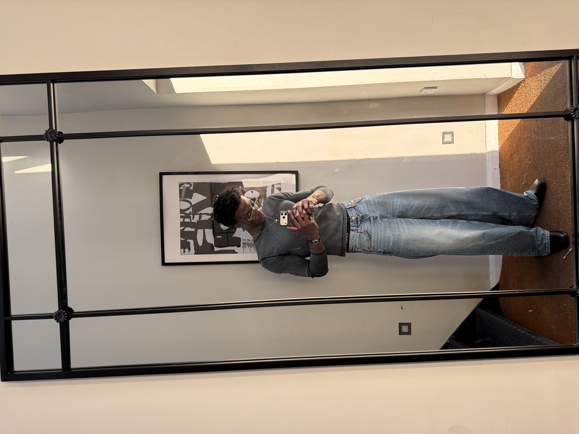 A mirror selfie taken in a room. A Black male stands in front of the mirror, facing it. He is wearing glasses, a long-sleeved grey shirt, blue jeans, and black shoes. He is holding a smartphone in his hands and taking a photo. The mirror is a grid of panes with a black frame. The room has a wall with a black and white framed print. A stairwell and a brown floor are visible in the mirror reflection. The location is likely a flat or an apartment. The city of London, United Kingdom cannot be confidently determined from the image.