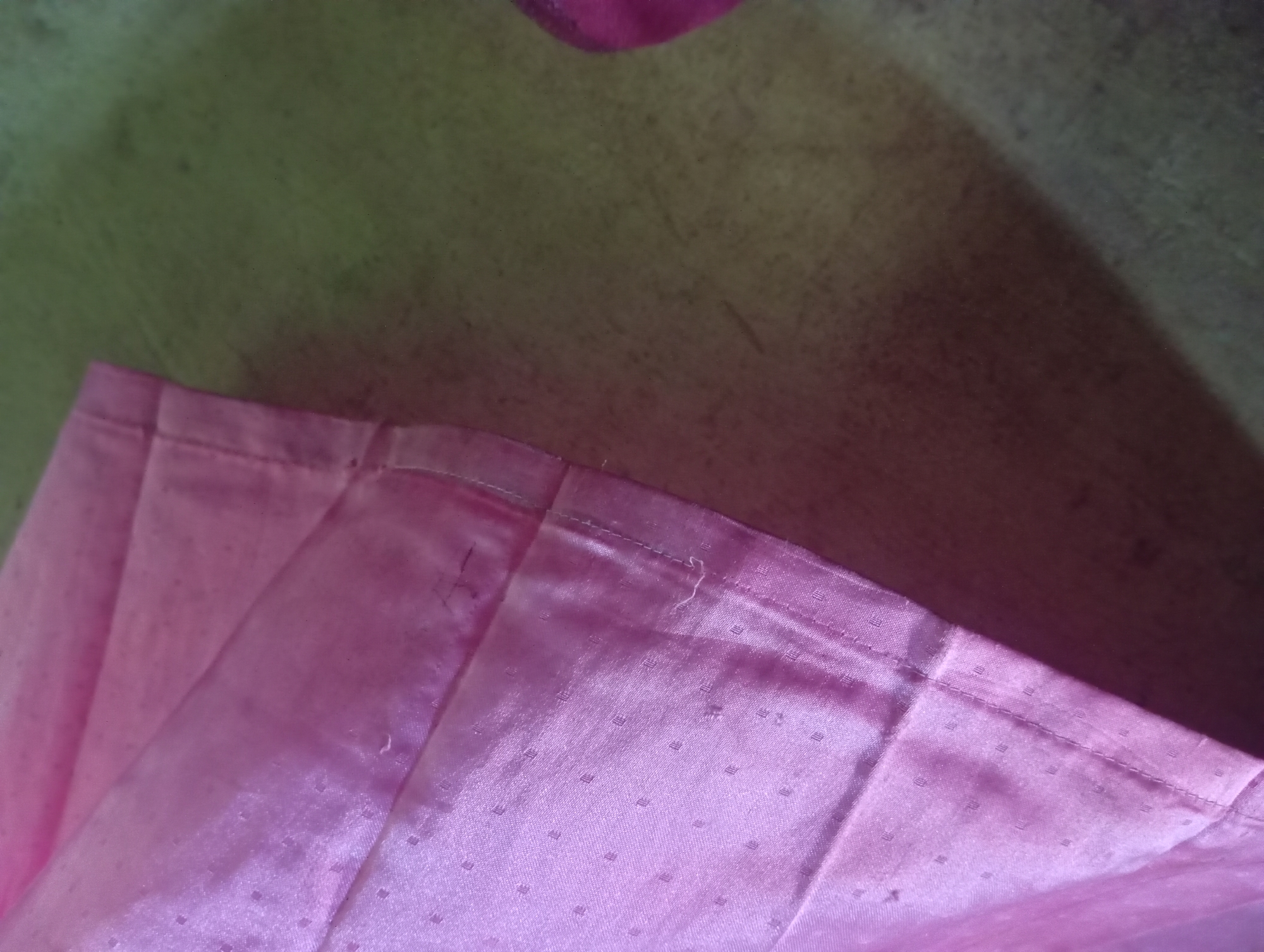 A close-up, eye-level shot shows a folded pink fabric with a subtle textured pattern of tiny squares. The fabric appears to be part of a garment or drapery, with visible stitching along a hemline. The texture of the fabric is smooth and slightly lustrous, reflecting light in soft highlights. The lower portion of the image shows a blurred surface, possibly a table or floor, with a textured grain and varying shades of muted green and brown. A hint of a darker pink object is visible in the bottom center, out of focus. The scene suggests an indoor setting, possibly domestic. There is no discernible evidence of activity or interaction within the frame. The image does not contain any elements that can be specifically identified with the city of Rejosari, Indonesia.