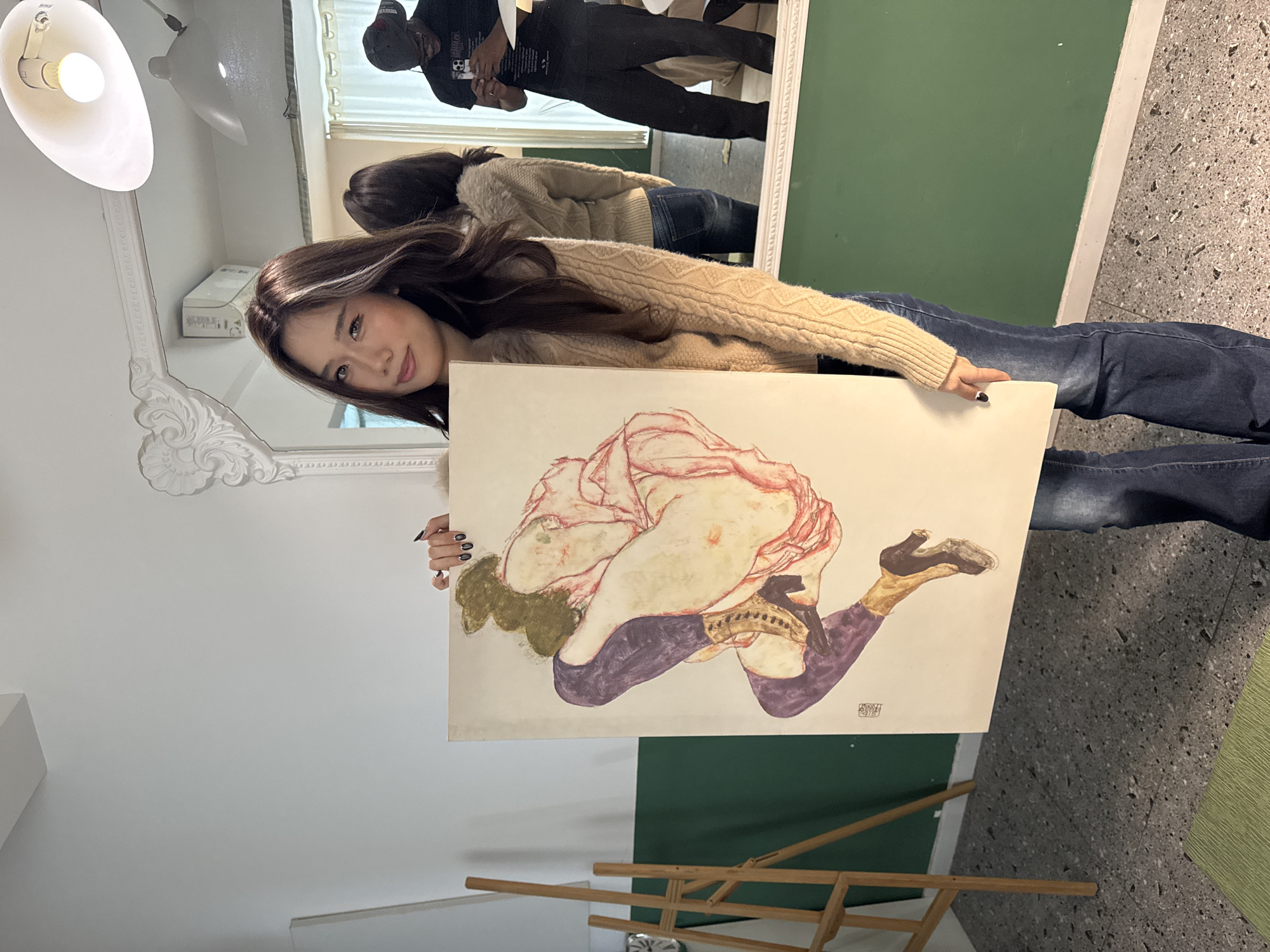 An East Asian female is depicted standing indoors in Liangjiang, China, holding a vertical rectangular artwork. The female, smiling and looking towards the camera, wears a beige knitted sweater with a faux fur collar and dark blue jeans. Her fingernails are painted black. The artwork she holds appears to be a reproduction of a piece in the style of Egon Schiele, depicting a crouched or seated female figure with dark hair, wearing purple leggings/stockings and brown heeled boots. A small dark mark, possibly a signature or stamp, is visible in the lower left of the artwork.
The environment features white upper walls and dark green lower walls. A large, ornate white-framed mirror dominates the right side of the background. Reflected in this mirror is a dark-skinned male wearing a black t-shirt and a backward black baseball cap, actively looking at a white smartphone held in both hands. An air conditioning unit is mounted above the mirror. Overhead lighting fixtures, including a bare bulb and a large white conical pendant, are visible. The floor is composed of dark gray terrazzo. A wooden easel and a white canvas are partially visible on the left side of the scene. A light green textured object rests on the floor in the lower left.