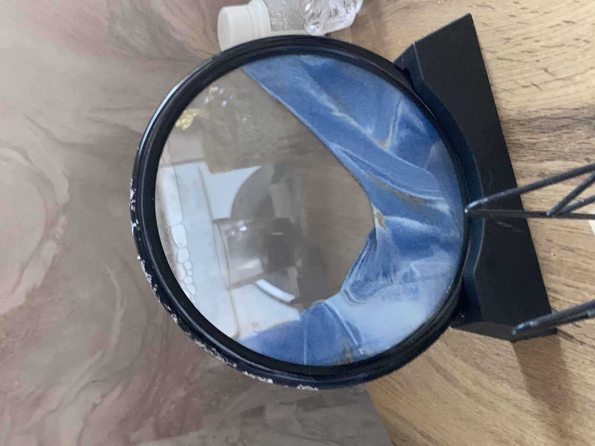 The image presents an overhead view of a circular object with a black frame, resembling a magnifying glass or a decorative mirror, resting on a black stand. The frame of the circular object shows some white speckles or wear. Within the circular object, reflections reveal a vibrant blue fabric, possibly denim or a similar textile, with faint gold-colored streaks or accents.
The object is placed on a two-toned surface. To the right, a light-colored wooden surface with a visible grain pattern serves as the base. To the left and in the background, a lighter, more abstract surface with swirling, marble-like patterns is visible.
In the upper background, slightly out of focus, are a small white bottle or cap with indistinct text and a clear, textured container, likely made of glass or plastic. The black stand supporting the circular object has both a solid rectangular component and thin, wiry supports. No people are visible, and no specific activity is taking place; the image is a still life captured indoors. There are no discernible cues within the image to suggest the time of day or the location of Stavropol, Russia.