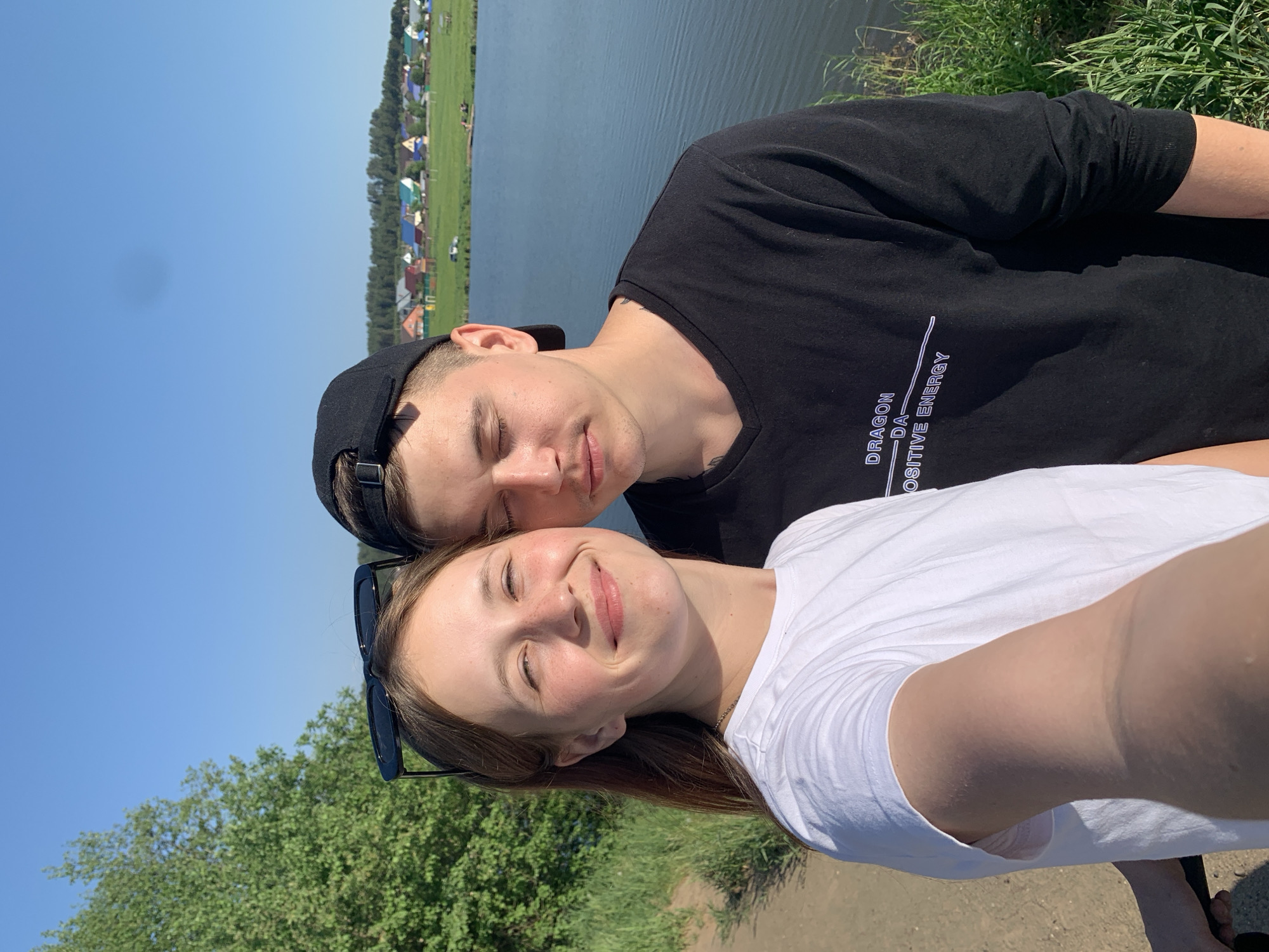 A selfie shows a young man and woman standing near a body of water on a sunny day. The man wears a black t-shirt with white writing, and a black baseball cap. The woman wears a white t-shirt and has sunglasses pushed down. They are both smiling and have their eyes closed. A body of water with houses on the other side is in the background. Green foliage and a dirt path are visible. The scene is likely outdoors near a waterfront area in Фоки, Russia.
