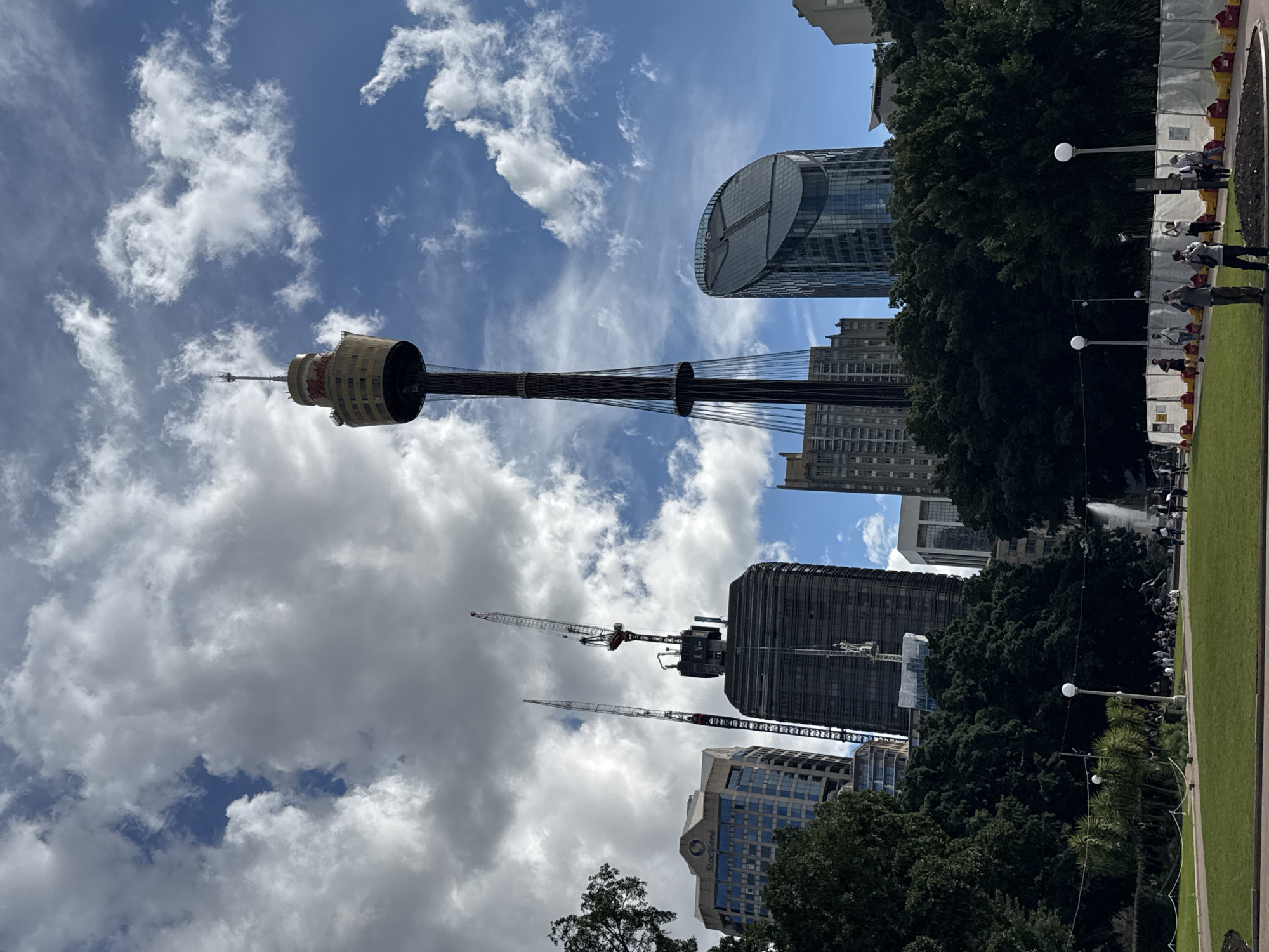 In an urban park setting under a partly cloudy sky, a tall observation tower dominates the background, distinguished by its round observation deck. The tower is flanked by several high-rise buildings, including one with a curved glass facade and another under construction with prominent cranes. In the foreground, a lush green lawn stretches towards a pathway lined with trees. Several people are scattered across the park, some walking, others sitting, and a few are engaged in taking photographs. Further into the park, a fountain can be seen. A construction barrier with red and yellow markings is also visible along the edge of the park. The scene captures a day in a city park with a mix of recreational activities and ongoing urban development.