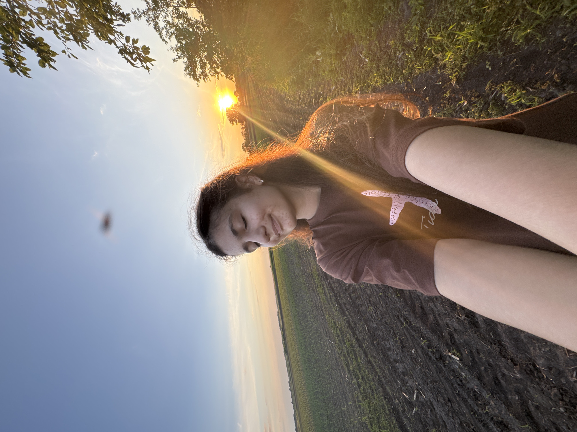 This image captures a young female at sunset in Ohuivka, Ukraine. The subject, positioned centrally in the foreground, has long, dark hair and wears a brown short-sleeved t-shirt with a light-colored graphic. Her eyes are closed, and her mouth displays a slight upward curve, consistent with a smile. Her face and hair are strongly backlit and illuminated by the warm, intense light of the setting sun, originating from the right of the frame. Her extended forearms are visible in the lower foreground, consistent with the pose for a selfie.
The background on the left features a vast agricultural field with visible rows of nascent green crops against dark, tilled soil, extending towards a distant horizon. The sky above the field transitions from clear blue at the top-left to hues of orange and yellow towards the sunset on the right. On the right side of the image, a narrow dirt path or track runs parallel to the field, bordered by green vegetation and sparse trees. A bright, circular sun disc is visible on the right horizon, producing significant lens flare across the scene. A blurred, dark, irregularly shaped object is visible in the upper-central portion of the sky, indicative of an object in close proximity to the camera lens.