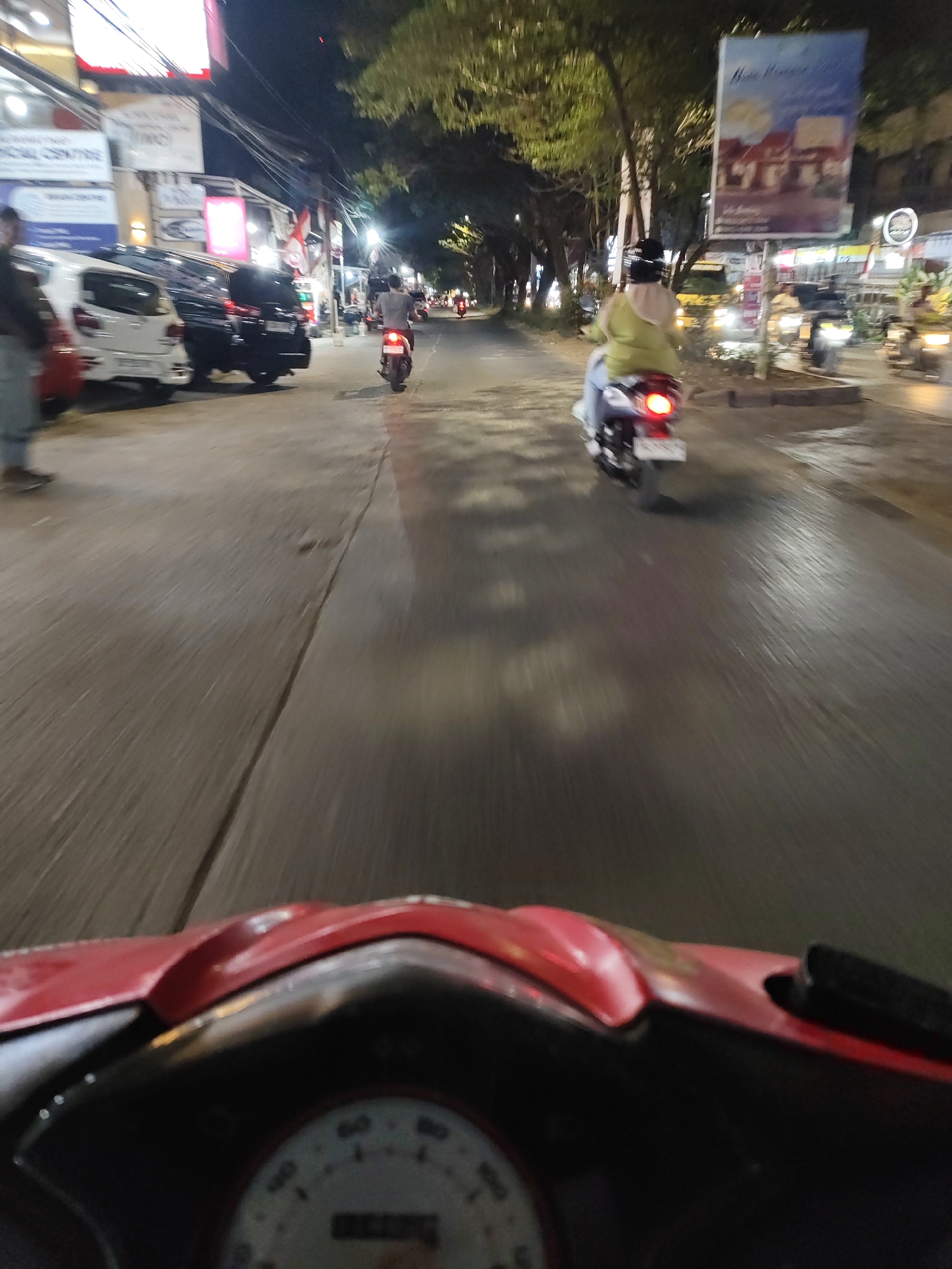 The image depicts a street scene at night, likely in Makassar, Indonesia, based on the context provided. The foreground is dominated by the handlebars and speedometer of a red motorcycle, indicating a first-person perspective from the rider. The road surface is wet, reflecting the artificial lights from streetlamps and buildings.
In the mid-ground, several vehicles are visible, including parked cars (one white, one black) and motorcycles in motion. A person on a white motorcycle with a black helmet and a lime green jacket is riding away from the camera. Another person on a red motorcycle is further down the road, also moving away.
The background shows a row of buildings with illuminated signs and awnings. Trees line the sidewalk, their leaves illuminated by the streetlights. A large billboard with an image of houses is prominent on the right side of the street. The overall impression is of a busy urban street at night, with traffic and commercial activity.