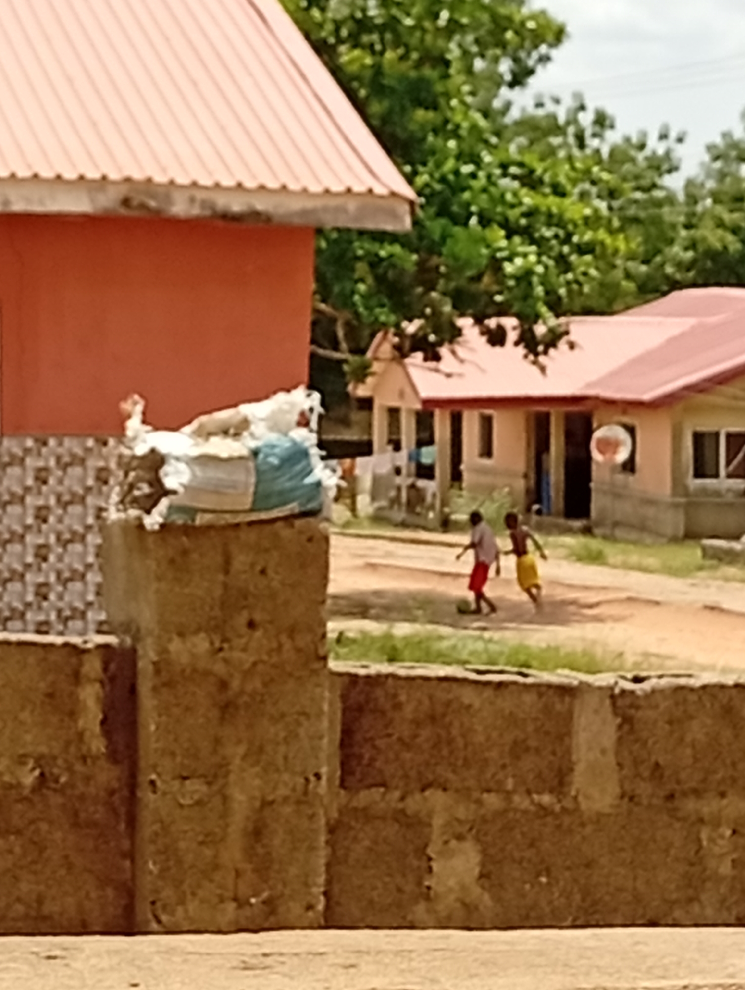 The image captures an outdoor scene in Adankolo, Nigeria, featuring two children playing in a dirt area in front of residential buildings. The time of day appears to be bright daytime, under a clear or partly cloudy sky, with ample sunlight.
In the midground, two young children, possibly boys, are engaged in playing what looks like soccer or football. One child wears red shorts and a light-colored top, while the other wears yellow shorts and a dark-colored top. A light-colored, spherical object, likely a ball, is near their feet. They are in motion, suggesting active play.
The background consists of several single-story houses with reddish-orange corrugated metal roofs and light-colored, possibly beige or pinkish, painted walls. One house further back appears to have a basketball hoop-like structure attached to its side. Lush green trees and foliage are visible behind and between the houses, indicating a green environment.
In the immediate foreground, to the left, is a concrete block structure or wall, topped with a white and blue bag that appears to be full, possibly of refuse or goods. Another concrete block wall runs across the bottom of the frame. A portion of a building with a red roof and a decorative tiled pattern on its lower wall is visible on the far left. The ground where the children are playing is unpaved, dusty earth. No visible text is present in the image.