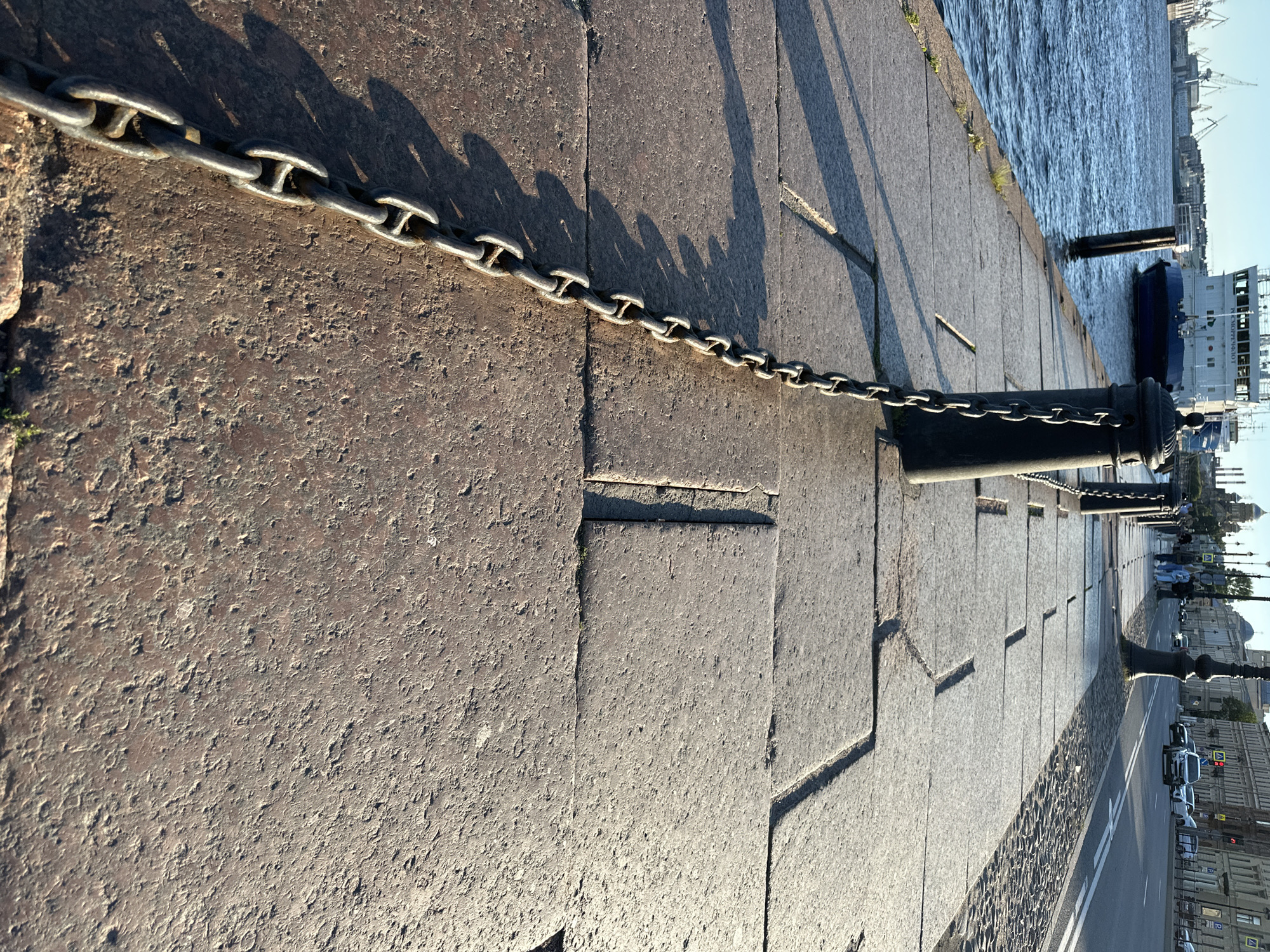 The image shows a paved embankment along a body of water in Saint Petersburg, Russia. A large metal chain stretches diagonally across the foreground, secured to bollards along the embankment. In the background, a ship is docked near the shore, and a street with cars and pedestrians runs parallel to the water. The architecture visible suggests a historic urban setting. The lighting indicates it is either early morning or late afternoon, with long shadows cast by the bollards and chain on the stone pavement.