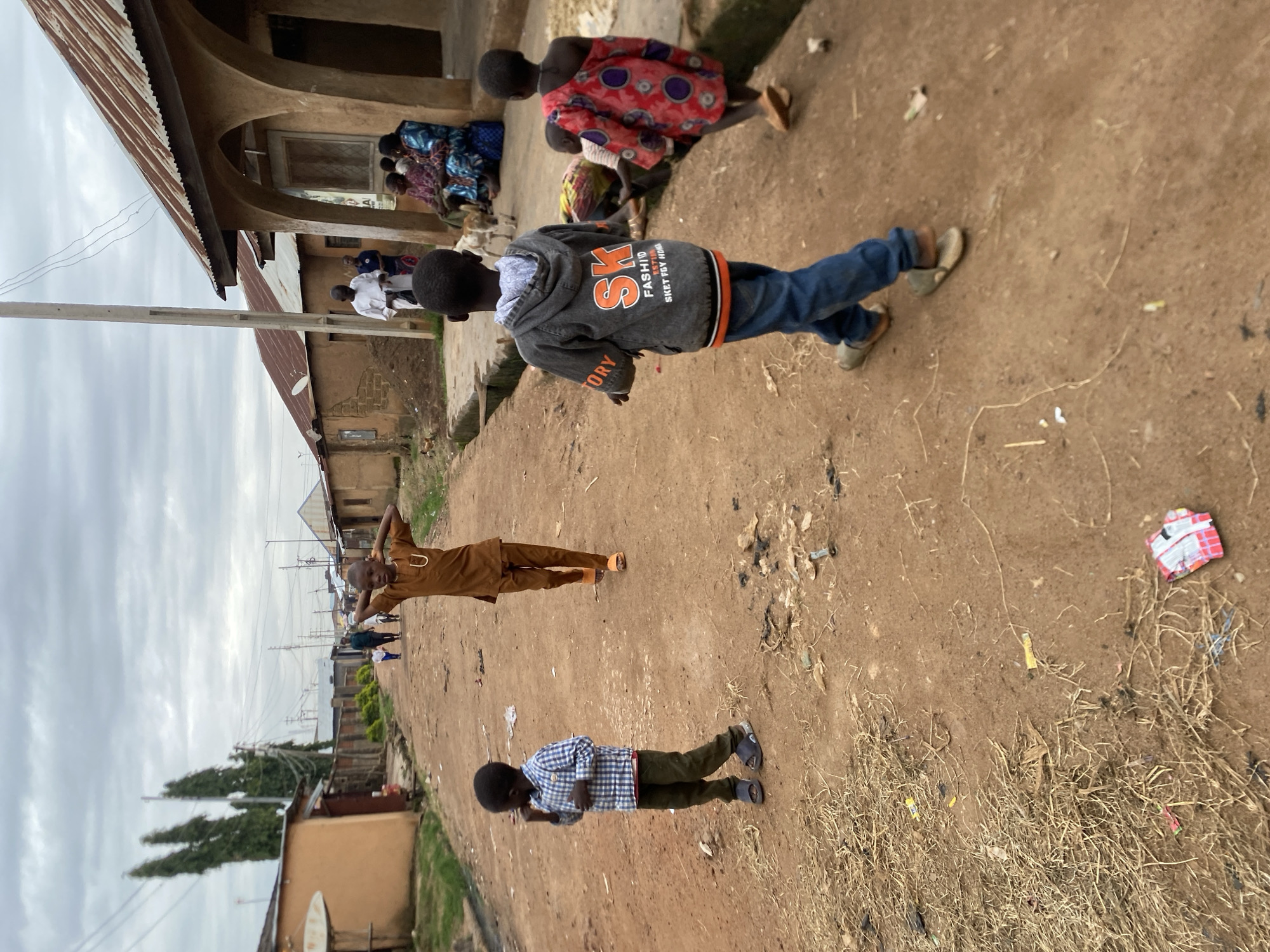 A group of children are walking down a dirt road in Bukuru, Nigeria. The road is lined with simple, mud-brick buildings with corrugated metal roofs. The sky is overcast.
In the foreground, a young boy in a blue and white checkered shirt and green pants is walking away from the camera. To his right, a boy in a brown outfit with a U-shaped design on the chest is stretching his arms above his head. Further down the road, two more children are visible. One is wearing a gray hooded jacket with "SK FASHION" printed on the back, and blue jeans. The other is wearing a red and white patterned dress.
In the background, several adults are gathered in front of one of the buildings. One man in a white robe is holding a white bag. A woman in a blue and patterned outfit is sitting with other individuals. A few dogs are visible in the vicinity. The ground is littered with dry grass and debris. Power lines crisscross the sky.