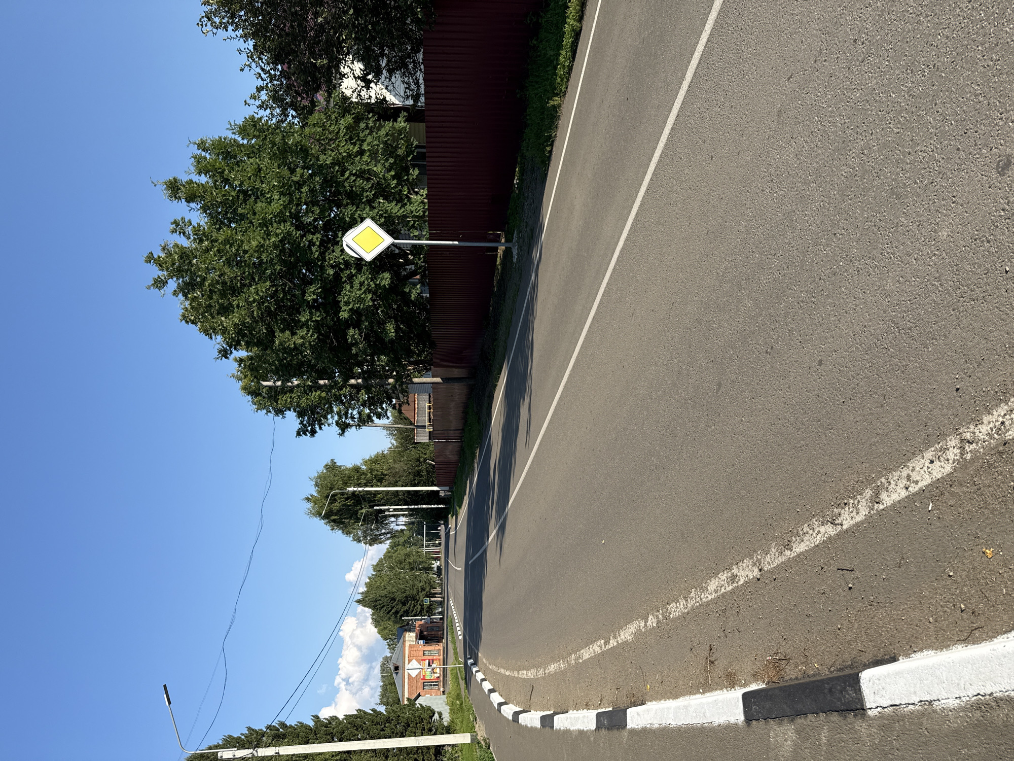 The image depicts a paved road in a rural or suburban setting, possibly in Krasnogorskoye, Russia, under a clear blue sky. The road curves to the left and recedes into the distance. On the left side of the road, there is a sidewalk with a white and black curb. Trees line the sides of the road, with a red corrugated metal fence visible on the right. A yellow diamond-shaped road sign with a white border is positioned on the right side of the road, likely indicating a priority road. A few buildings, including a red brick structure, are visible in the background on the left. Streetlights are present along the road. The environment appears to be daytime, with sunlight casting shadows. There are no people or animals visible in the image.