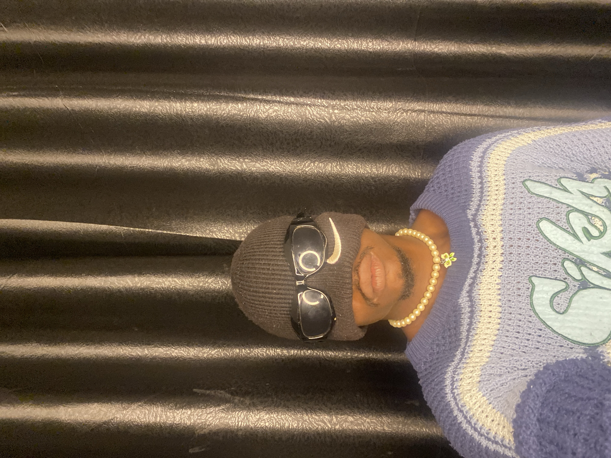 In Abakaliki, Nigeria, a person is photographed from the chest up, against a backdrop of dark, vertically pleated curtains. The individual is wearing a dark knit beanie that covers their forehead and extends down to the bridge of their nose, obscuring their eyes. Large, dark-rimmed sunglasses are perched on the beanie, positioned over where the eyes would be. A faint Nike swoosh logo is visible on the left lens of the sunglasses. Below the sunglasses, the person's nose and mouth are visible, along with a thin mustache and goatee. They are also wearing a pearl necklace with a small, green and yellow flower pendant. The person's upper body is adorned with a light blue, knitted sweater featuring horizontal stripes of white and a lighter shade of blue. The sweater has a textured, open-knit pattern and a large, light blue graphic design on the front that appears to read "Sikh". The lighting in the image is warm, casting shadows and highlighting the texture of the curtains and the clothing.