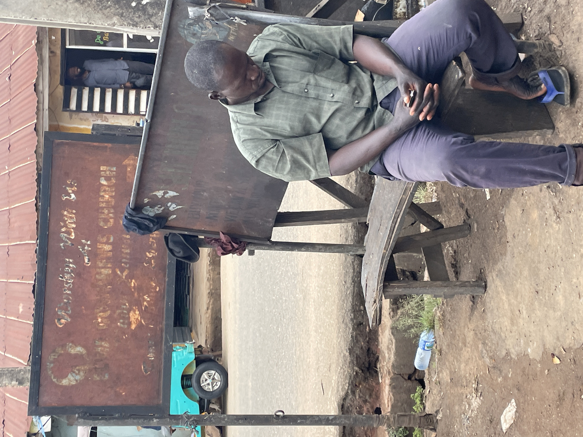 A man is seated on a weathered wooden bench, leaning slightly forward with his hands clasped in his lap. He is wearing a light green checkered button-down shirt and dark trousers, and he has sandals on his feet. To his left is a large, rectangular sign with faded or rusted writing, partially obscured. The sign's inscription includes the words "CHURCH" and possibly "Daughters Zion Women's Church." Behind the sign, a glimpse of another person is visible inside a building with a barred window. In the background is an exposed road surface. The setting appears to be an urban area, possibly the city of Jos, Nigeria. Litter, including a plastic water bottle, lies on the ground near the bench. A turquoise three-wheeled vehicle is partially visible behind the sign.