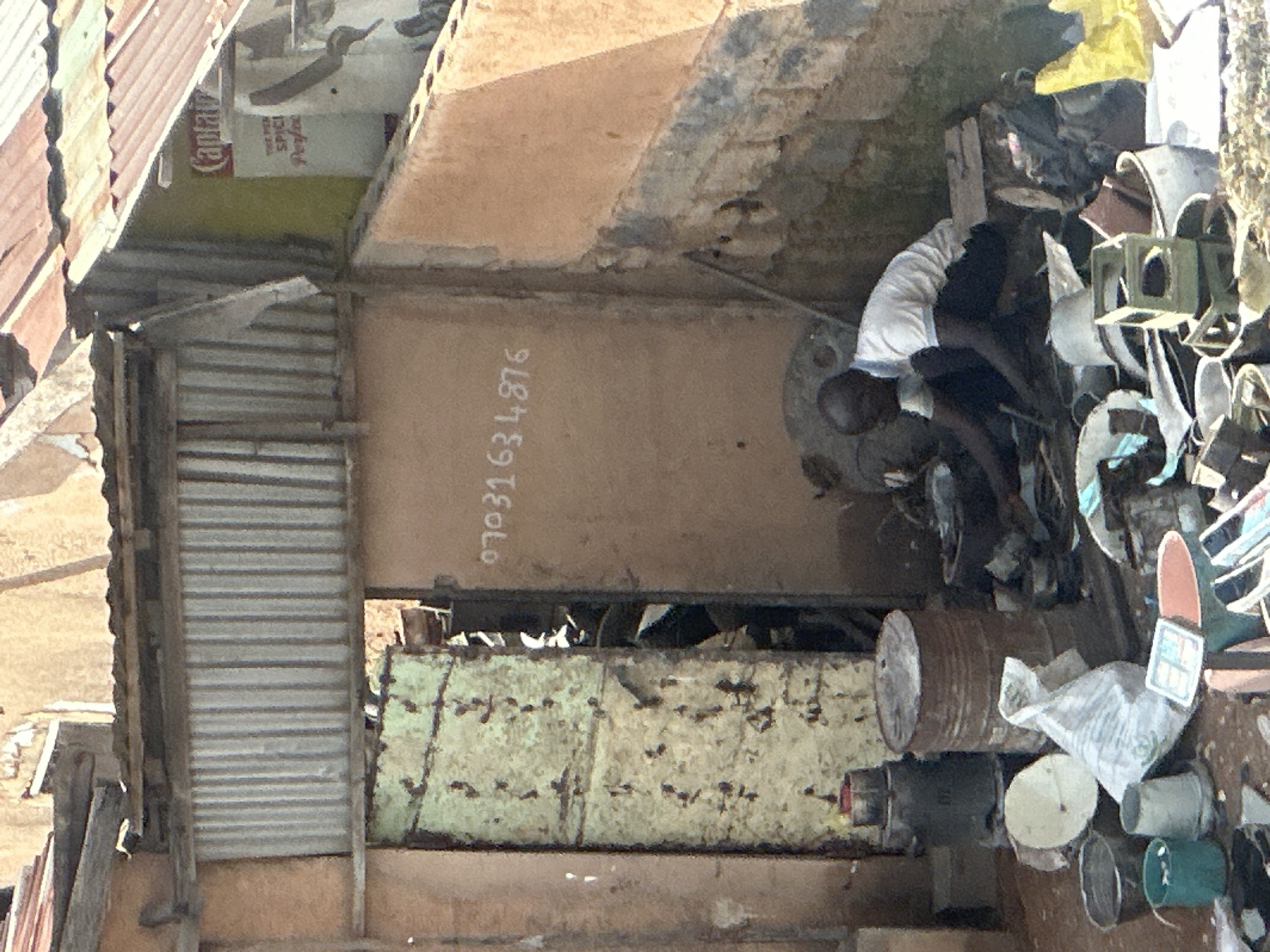 A man is working in a cluttered outdoor area in Jos, Nigeria, surrounded by various objects. The scene appears to be a workshop or storage area, possibly for scrap materials. In the background, a corrugated metal roof extends over part of the scene, with a partial view of a building constructed with what appears to be corrugated metal and possibly cinder blocks. A sign with "Captain" and "SPICI" is visible on the upper right.
The man, wearing a white polo shirt and dark pants, is crouched down, focusing on something in front of him amongst the scattered items. These items include several metal barrels, buckets, plastic containers, a white plastic bag, and other unidentifiable debris. The ground is covered with dirt and more assorted objects. An open doorway, with a weathered, green-painted door leaning to the side, reveals more clutter within the interior. A phone number, "07031634876", is scrawled in white on the wall to the right of the doorway. The overall impression is one of a busy, informal workspace characterized by a high degree of disarray.