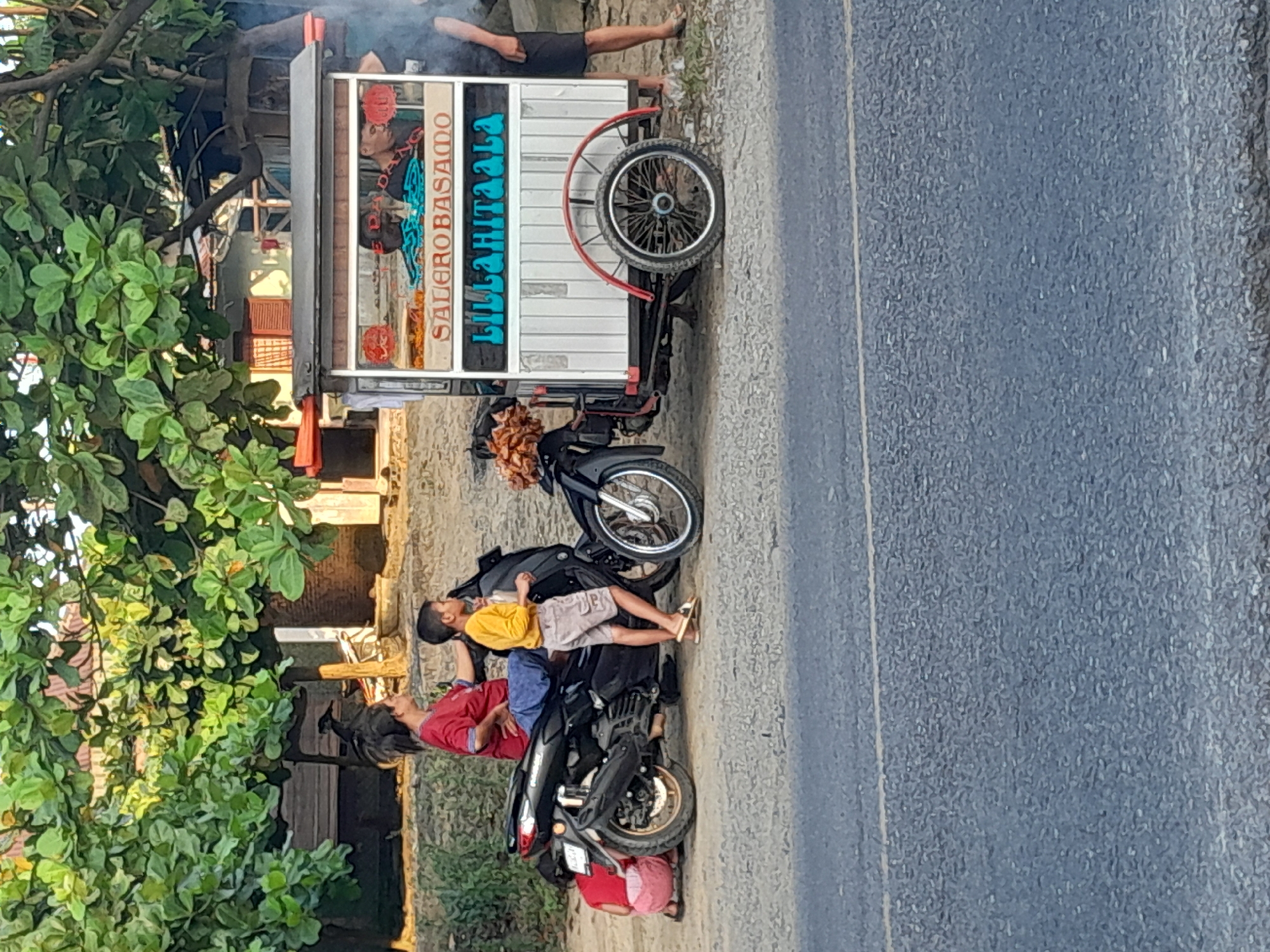In Pekan Tolan, Indonesia, a street scene features a food cart adorned with the text "SALEROBASAMO" and "LILLAHITAALA". The cart is attached to a tricycle and appears to be selling food. Several people are present. A woman in a red shirt sits on a motorcycle. A young boy in a yellow shirt stands beside the motorcycle. Another child, wearing a pink garment, is crouched near the rear of the motorcycle. Two men are partially visible behind the food cart. The background includes trees and buildings. The foreground is a paved road.