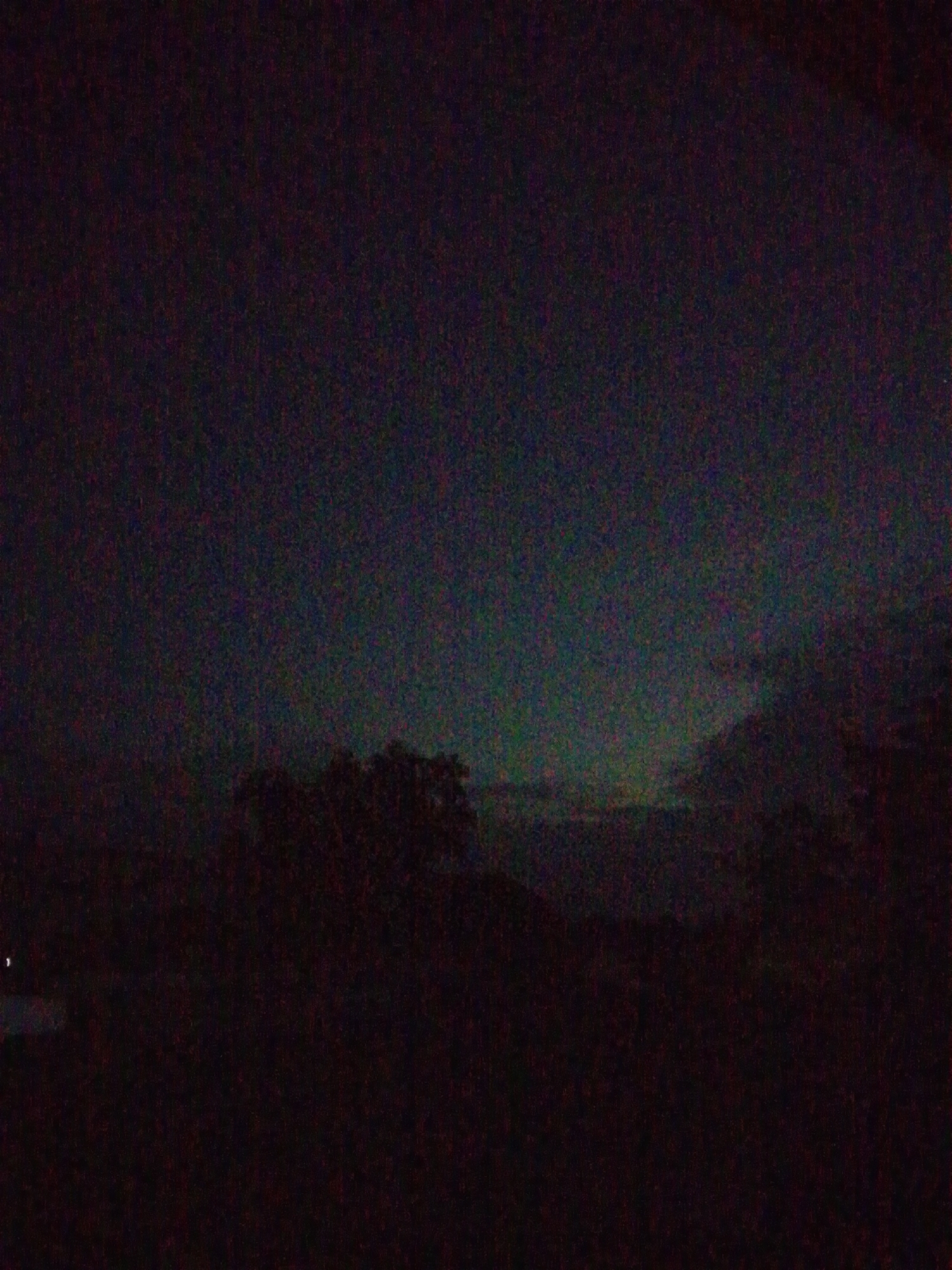 The image depicts a nighttime sky over a dark, silhouetted landscape, likely in or near Serti, Nigeria. The sky exhibits a faint greenish-blue aurora-like glow, a phenomenon not typically observed at this latitude, suggesting a possible atmospheric anomaly or an unusual display of the aurora borealis/australis. The lower portion of the image is dominated by dark silhouettes of trees and possibly some low-lying vegetation. There is a faint point of light on the far left, which could be a distant artificial light source. The overall impression is one of a vast, dark expanse with a subtle, ethereal glow in the upper atmosphere. The image is grainy, consistent with low-light photography.