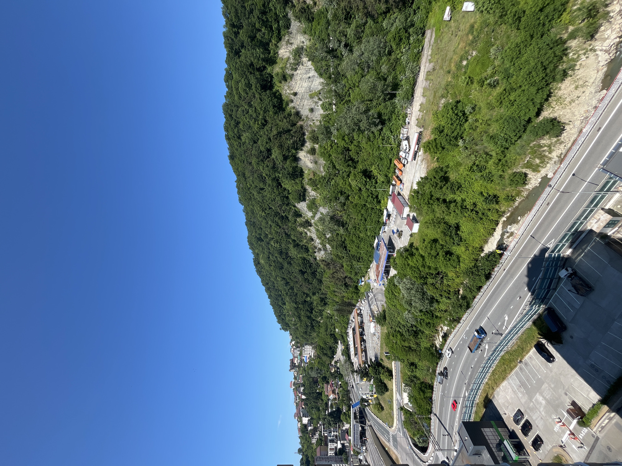The image captures an elevated, panoramic view of a highway winding through a lush, green mountainside in Sochi, Russia. The clear blue sky dominates the upper portion of the frame, with a vibrant green forest covering the steep incline of the mountain. Several buildings, including a residential area with multi-story houses and a gas station with fuel pumps, are nestled amongst the trees and along the roadside. A multi-lane highway is clearly visible, with several vehicles, including cars, a truck, and what appears to be a bus, in motion. A parking lot filled with cars is also present in the foreground. The landscape appears to be a mix of natural beauty and urban development.