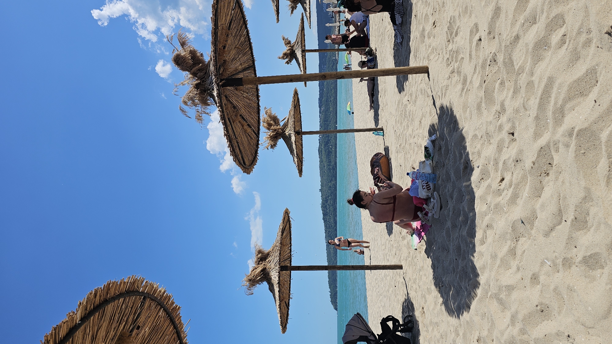 The image shows a beach scene in Varna, Bulgaria. Several straw umbrellas are positioned along the sandy shore, providing shade. A woman in a bikini sits on the beach, appearing to use a mobile phone. Personal belongings, including bottles of water, clothing, and shoes, are scattered around her. Further down the beach, other people are relaxing and walking along the shoreline. The blue sea is visible, meeting the horizon under a bright, clear sky with scattered clouds. A stroller is visible in the foreground.