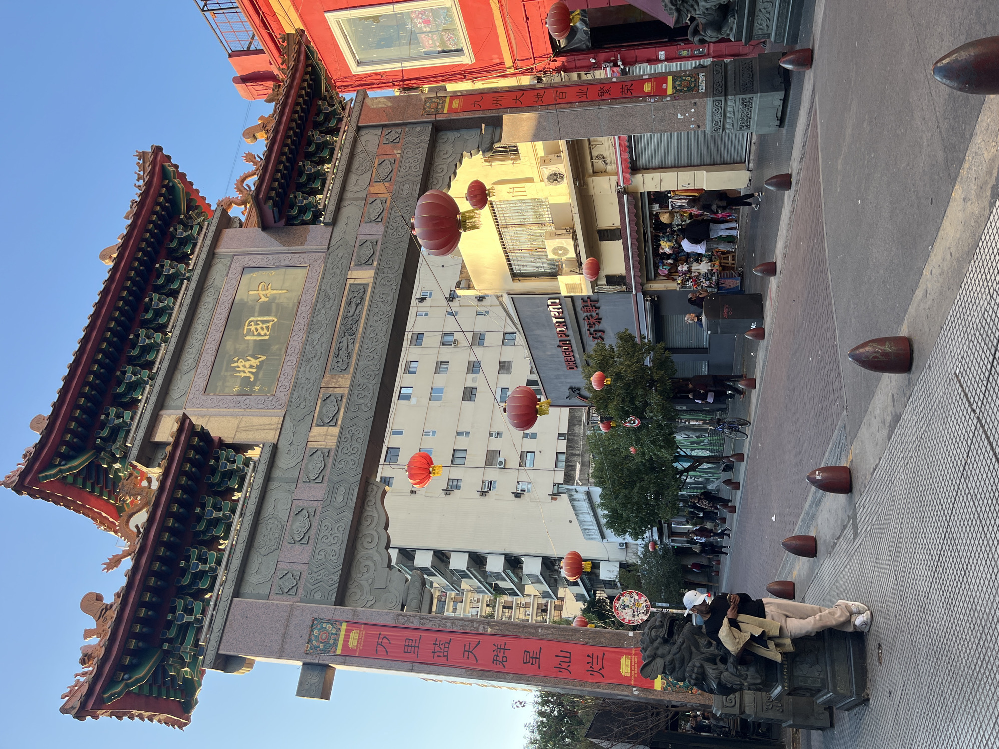 The image captures the ornate gate of Chinatown in Buenos Aires, Argentina, with a man resting on a stone base at the gate's base. The traditional Chinese gate is constructed from stone and wood and is adorned with intricate carvings, Chinese characters, and red lanterns. Buildings line the street, with pedestrians visible in the background.