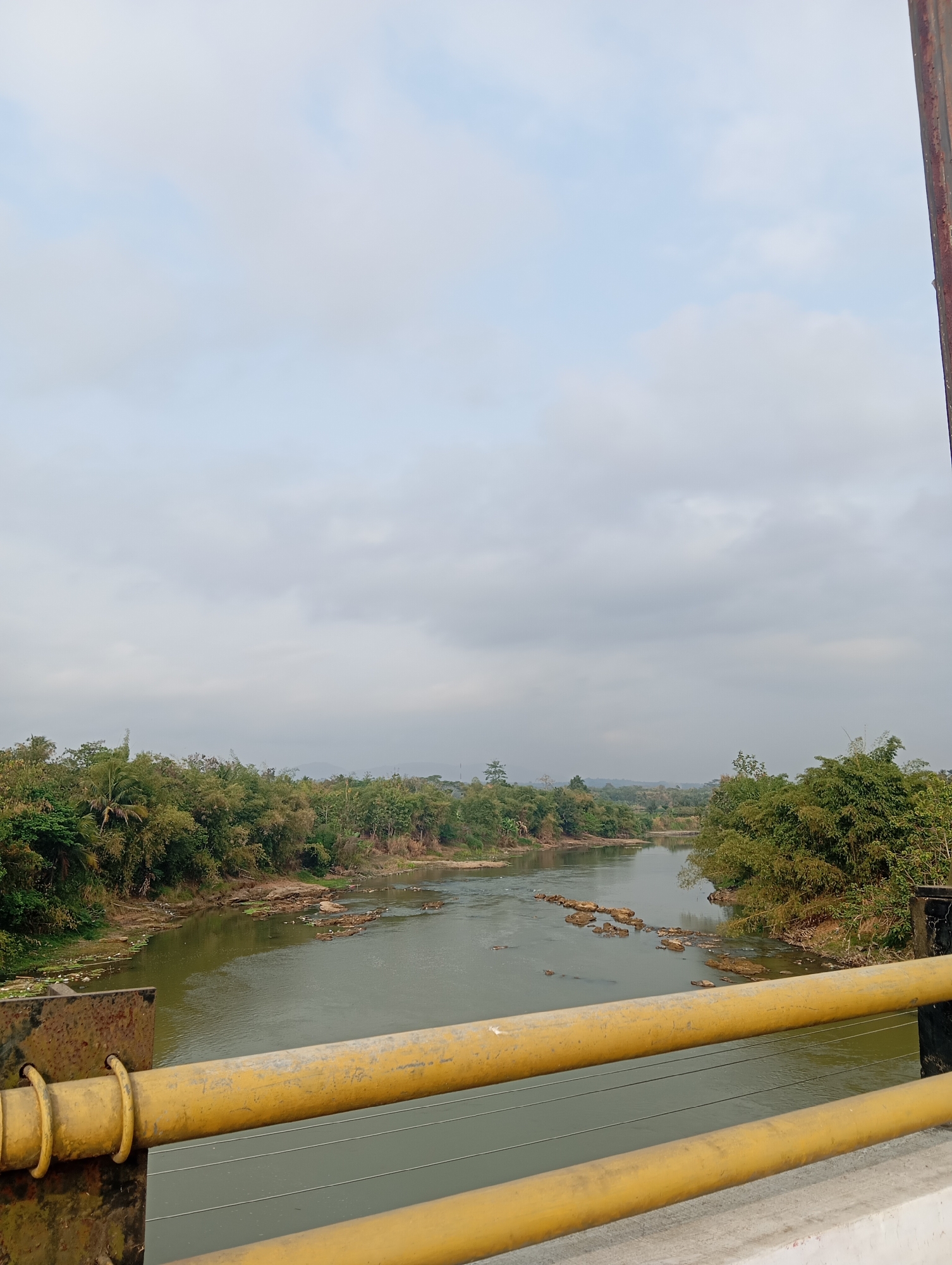 The image depicts a serene river scene in Kedungbenda, Indonesia, viewed from a bridge. The river, with its calm, green waters, meanders through a lush landscape of dense vegetation, including bamboo trees and other tropical foliage that lines its banks. Small clusters of rocks are visible protruding from the water, creating a natural, textured effect. The sky above is overcast with a muted, cloudy expanse, casting a soft, diffused light over the entire scene. In the foreground, the railing of the bridge, painted a bright yellow, creates a strong horizontal line, adding a man-made element to the otherwise natural environment. The presence of the bridge suggests a point of access or passage over the river, likely a common feature in the rural or semi-rural setting of Kedungbenda. The overall impression is one of tranquility and natural beauty, characteristic of a tropical riverine landscape.
