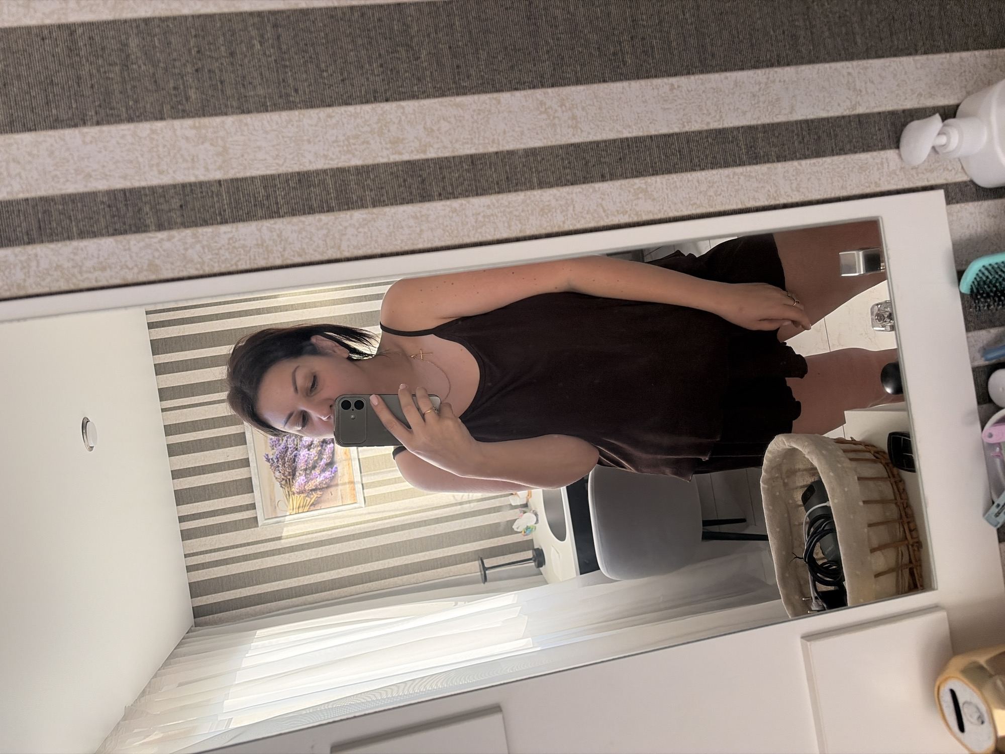 An image shows a woman taking a mirror selfie in an indoor setting, likely a bedroom or dressing area in Sochi, Russia. The photo is rotated, showing a woman with dark hair wearing a dark camisole and shorts, holding a dark-colored smartphone to capture her reflection in a large, white-framed mirror. A gold chain with a cross pendant is visible around her neck, and she wears rings on her fingers.
The reflection in the mirror reveals a room with prominent striped wallpaper or blinds on the far wall. A framed picture featuring purple lavender is hung on this wall. Natural light streams in through white blinds or curtains, suggesting it is daytime. A grey chair with a black base is visible to the right of the woman's reflection, next to a white surface that appears to be a dresser or vanity. On this surface, a woven basket with a fabric liner holds what looks like a hair dryer or another electronic device with a cord. A circular light fixture is visible on the ceiling in the reflection.
Outside the mirror frame, on the surface where the mirror rests, there is a light-colored striped rug or carpet. To the right of the mirror, a white pump bottle (possibly soap or lotion) and a light blue hairbrush are visible. Part of a gold-colored metallic object is also seen in the lower right corner. No discernible text is visible in the image.