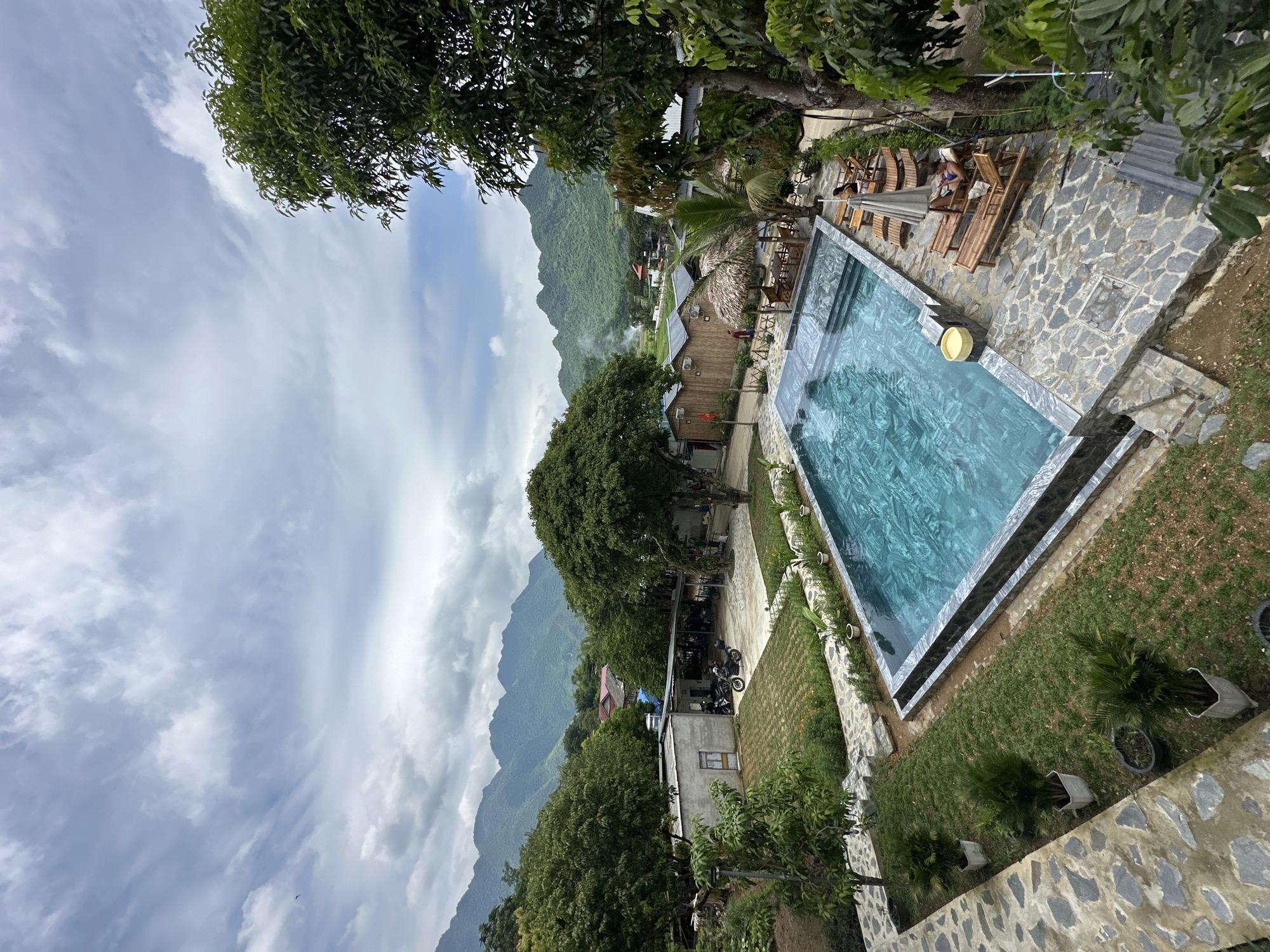 This is a high-angle exterior shot of a swimming pool area at what appears to be a resort or guesthouse in Xóm Khán, Vietnam. The pool is rectangular with blue-tiled water and dark gray stone coping and steps. Several lounge chairs made of bamboo are arranged around the pool, with two individuals visible relaxing on them. A thatched umbrella shades one of the chairs.
The surrounding environment features lush green grass, potted plants, and stone pathways. Beyond the pool area, there are several small wooden structures with thatched roofs, some appearing to be individual bungalows or cabins. In the middle ground, there are a few motorcycles parked near a simple, light-colored building.
The background is dominated by verdant, mist-shrouded mountains, characteristic of the Vietnamese landscape. The sky is overcast with a mix of light and dark clouds. The overall impression is one of a tranquil, semi-rural retreat surrounded by natural beauty.