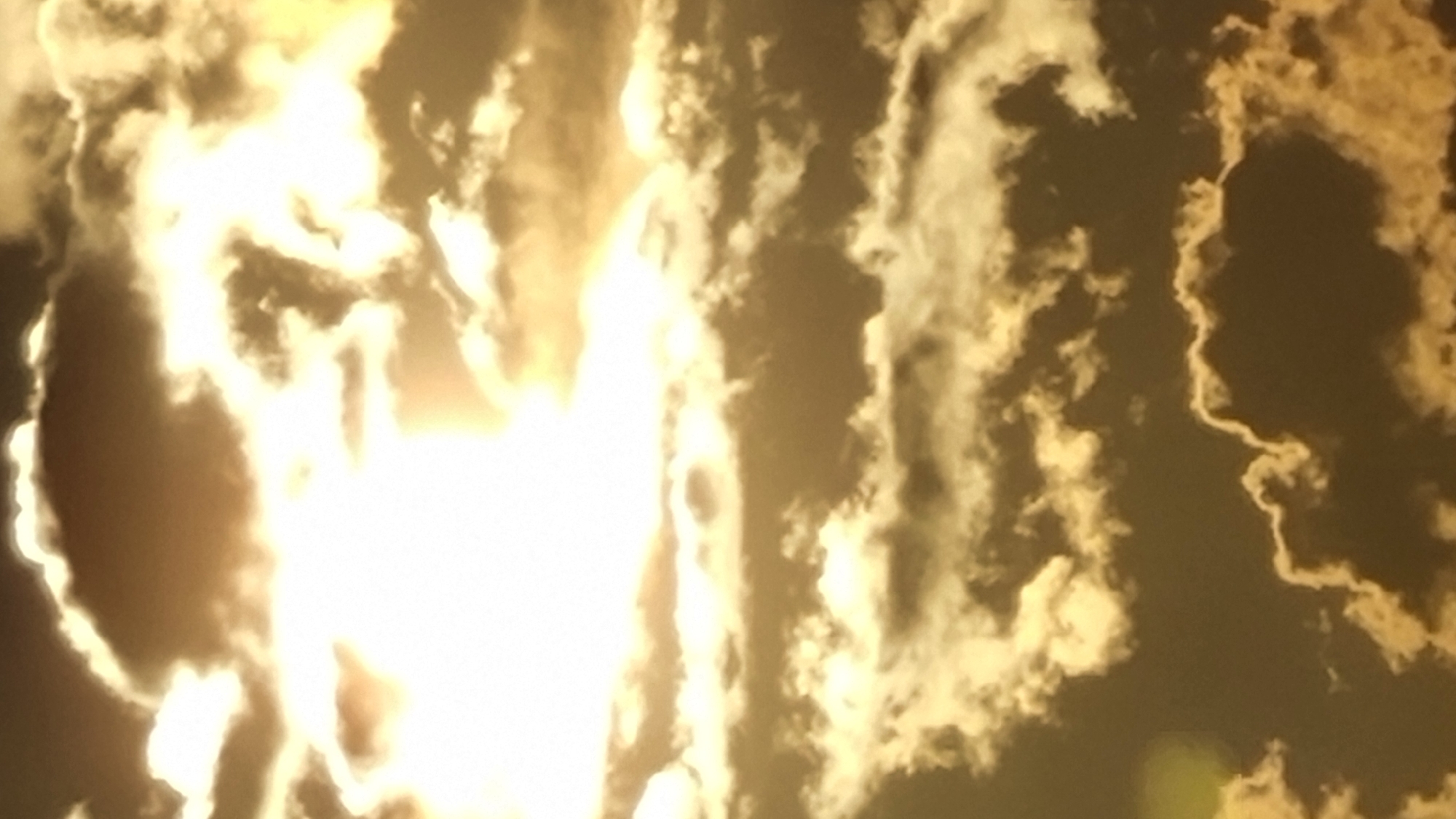 A low-angle vertical shot captures a dramatic sky filled with sunlit clouds. The sun, positioned in the upper left quadrant, is heavily obscured by dense, bright white clouds that are backlit by its intense light, creating a halo effect and a strong glow. The clouds vary in texture and density, with some appearing thick and dark, casting shadows, while others are wispy and illuminated. The overall color palette is dominated by golden hues, transitioning from bright yellows and whites in the sunlit areas to deep browns and grays in the shadowed regions. The bottom portion of the image shows more textured clouds with illuminated edges, some appearing to swirl and form intricate patterns against the darker sky. There are no people, structures, or discernible landmarks visible, suggesting this is a natural scene focused solely on the atmospheric conditions of the sky.