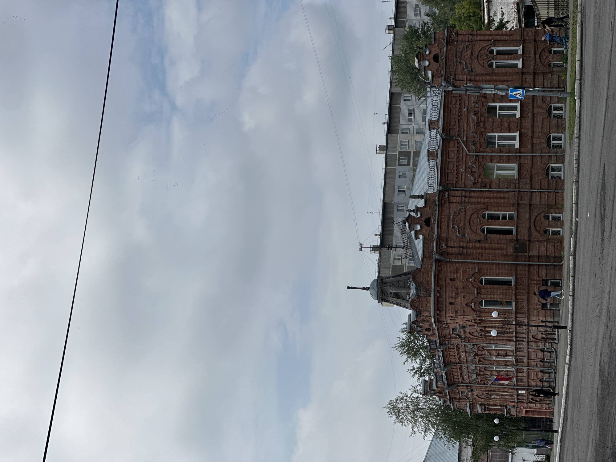 The image shows a large, ornate, two-story brick building with a complex roofline and multiple decorative elements. The building is made of red brick and has numerous windows, some arched. A Russian flag is visible on the front of the building. Several people are walking on the sidewalk in front of the building, and some are near the street. In the background, there is a multi-story apartment building and trees. The sky is overcast. The scene appears to be located in the city of Nogornyi, Russia.