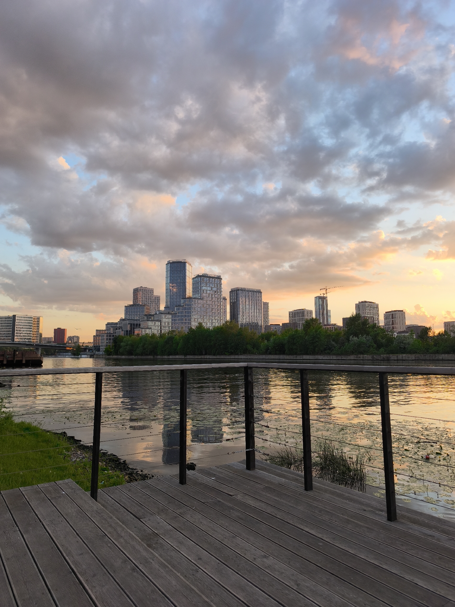 This captivating image showcases a modern cityscape in Moscow, Russia, at either sunrise or sunset, viewed from a wooden waterfront deck. In the immediate foreground, a dark wooden deck with horizontal planks leads towards a dark metal railing, featuring sturdy vertical posts and thin horizontal cables. To the left of the deck, a strip of green grass borders the water.
The midground is dominated by a calm body of water, likely a river, which acts as a mirror, reflecting the vibrant sky and the distant urban silhouette. Patches of aquatic plants, resembling lily pads, dot the water's surface, especially towards the right.
Across the water, the background reveals a striking skyline of contemporary high-rise buildings. These are mostly glass-fronted structures with sleek designs, varying in height and form, creating a dynamic architectural display. Some buildings exhibit terraced levels, while others are tall, rectangular towers. A dense line of green trees and foliage runs along the far bank, providing a natural buffer between the river and the urban development. On the far left, a bridge is visible spanning the water, leading to more buildings beyond. Towards the right, a tall construction crane rises prominently amidst the buildings, suggesting ongoing growth in the area.
The sky above is dramatic and dynamic, filled with an array of clouds. Darker grey clouds are interspersed with soft patches of blue sky, and are vividly illuminated with warm hues of orange, pink, and yellow along the horizon, characteristic of either dawn or dusk. These colors are beautifully mirrored in the water below. The scene is devoid of people or visible text, conveying a sense of serene urban beauty.
