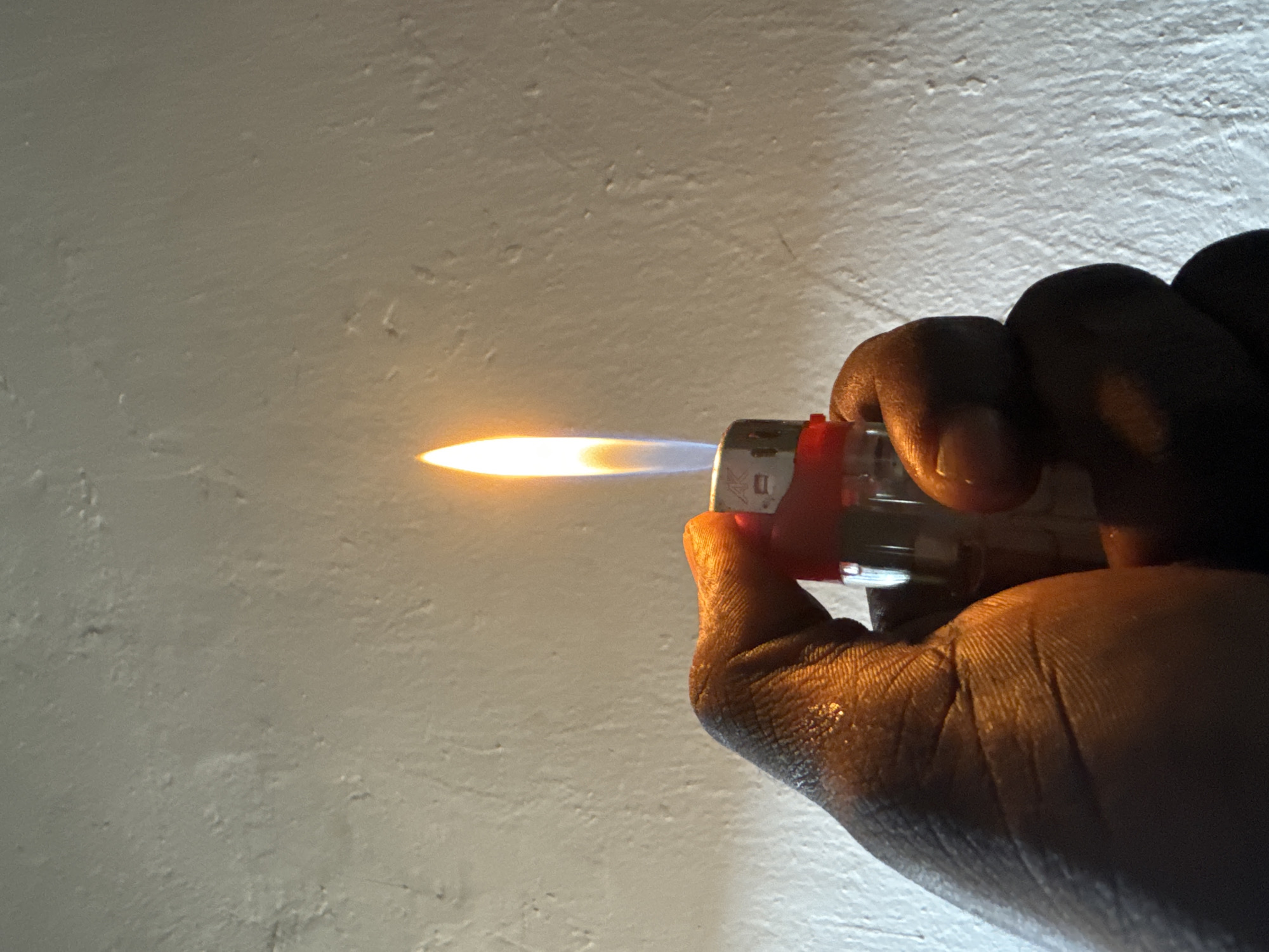 A close-up shot captures a hand holding a lit lighter. The lighter, predominantly clear with a red accent, emits a bright, orange flame with hints of blue at its base. The hand is dark-skinned and appears to be glistening, possibly from sweat or moisture. The background is a textured, off-white wall, showing subtle variations in color and surface. The lighting suggests an indoor environment, with the flame providing the primary light source for the scene. This image does not contain information to determine its location in Ifite-Awka, Nigeria.