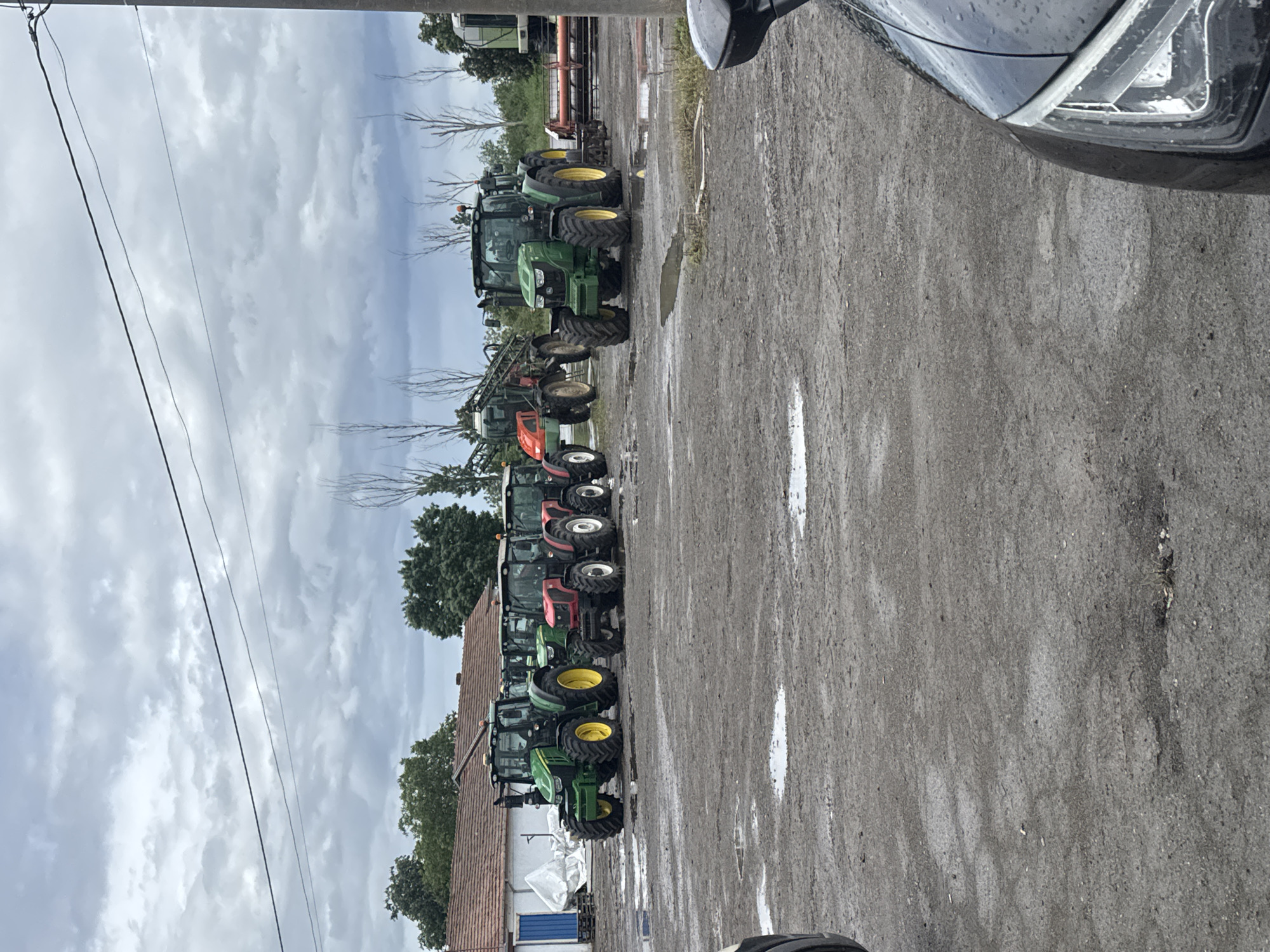 The image shows a row of tractors parked on a wet, paved surface. There are several green John Deere tractors, as well as some red and orange tractors. A building with a red tile roof and white walls is partially visible behind the tractors. Power lines run above the building. The sky is overcast. The front corner of a dark-colored car is visible on the right side of the image. The location is likely Sarnevo, Bulgaria.