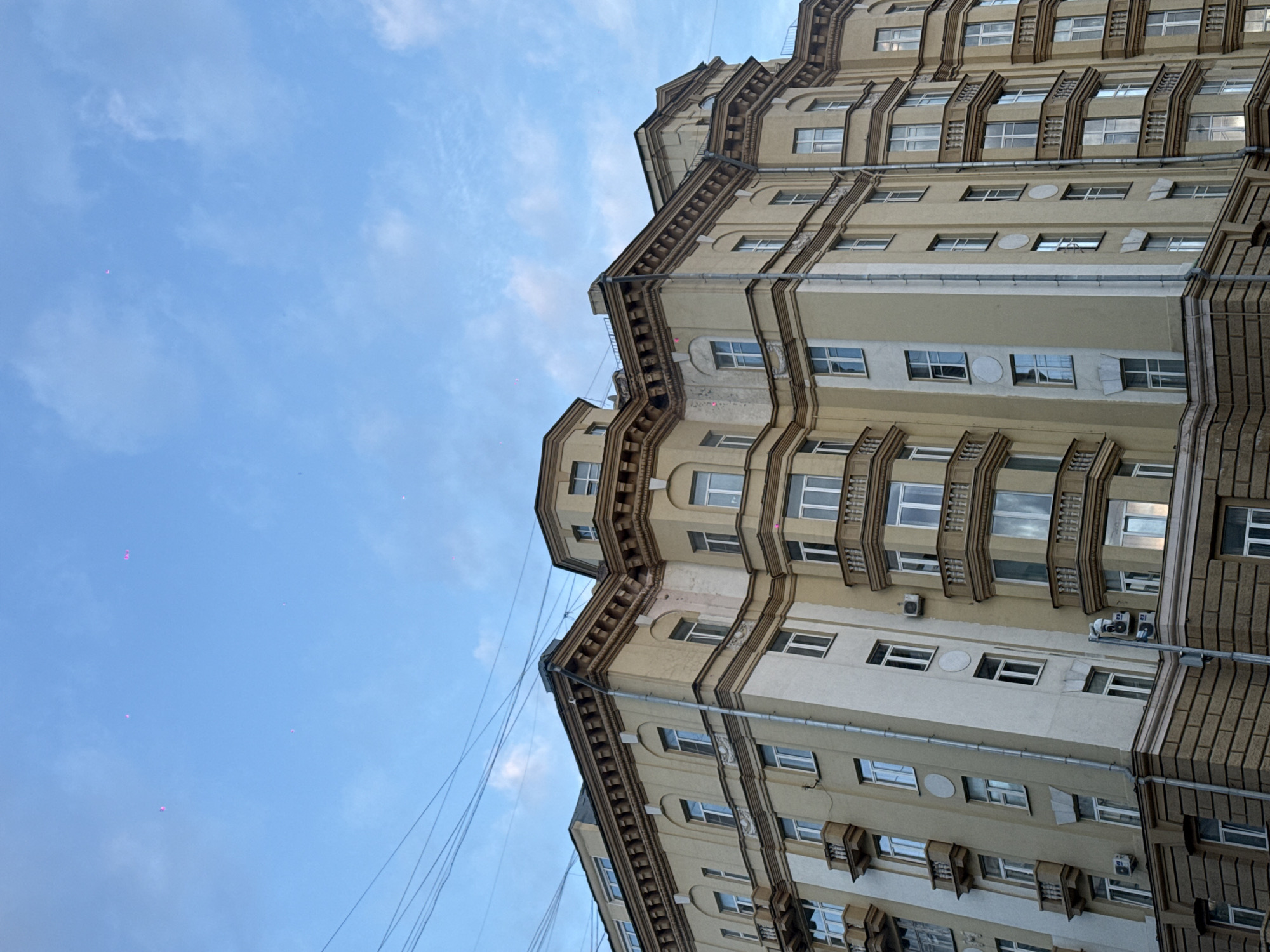 The image shows a tall, light-colored building with multiple floors in Moscow, Russia. The building's facade has decorative molding and multiple windows on each floor. The sky is blue with scattered clouds. Several thin wires run across the sky in the lower left of the image. No people are visible in the scene. The image is taken from a low angle, looking up at the building.
