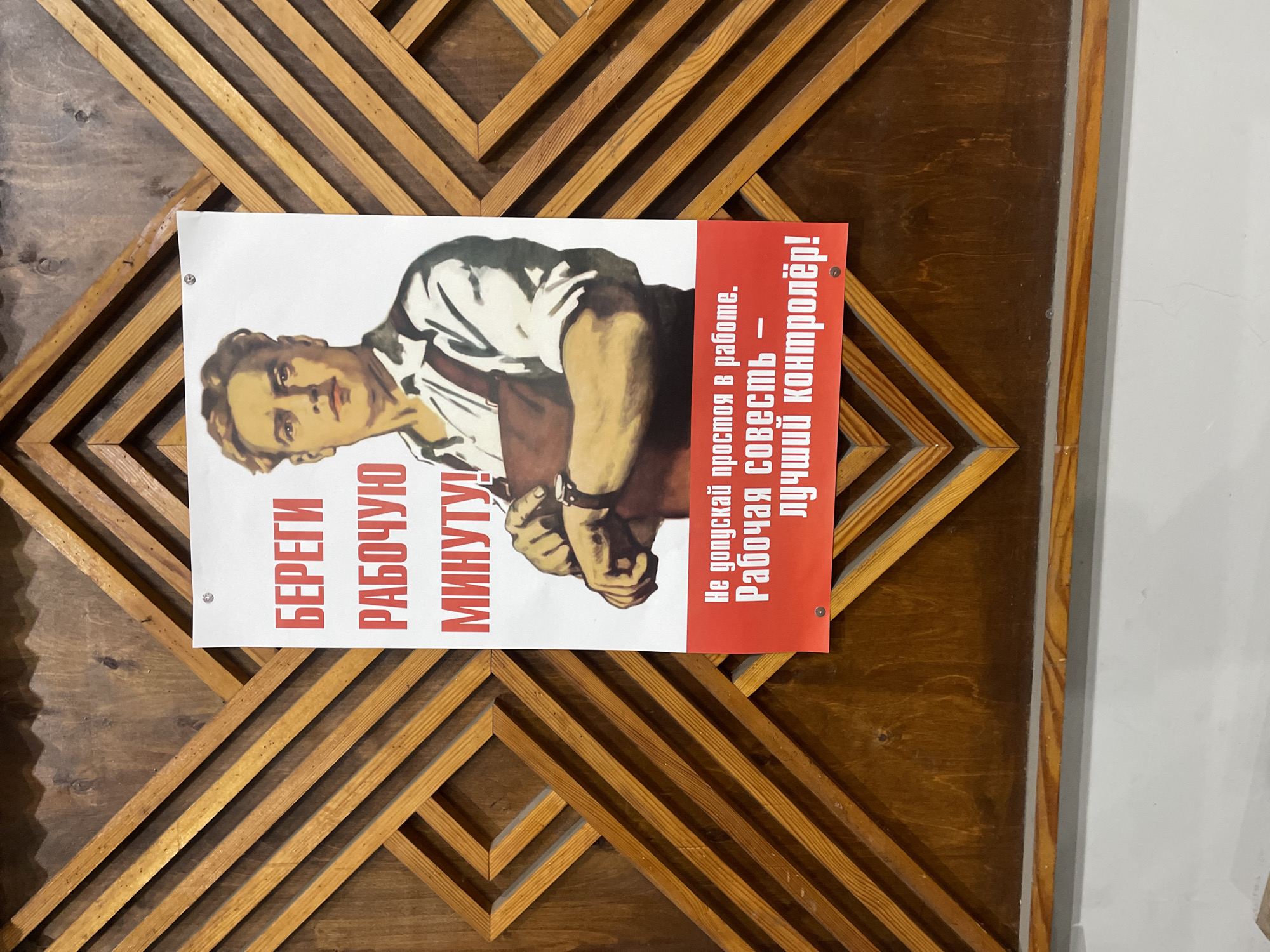 A propaganda-style poster featuring a serious-looking man is displayed on a wooden wall, likely in an indoor setting in Chernogolovka, Russia. The man, depicted from the chest up in a vintage artistic style, wears a light-colored shirt with rolled-up sleeves and has a determined expression. His hands are clasped in front of him, and he wears a watch on his left wrist, emphasizing the theme of time.
The poster conveys a message about workplace productivity and responsibility. On the left side, in large red capital letters, it reads: "БЕРЕГИ РАБОЧУЮ МИНУТУ!" which translates to "CHERISH EVERY WORKING MINUTE!" On the right side, against a red background, the text states: "Не допускай простоя в работе. Рабочая совесть — лучший контролёр!" This translates to "Do not allow downtime at work. Working conscience is the best controller!"
The poster is affixed to a wall that features a distinct geometric pattern of light wooden slats on the left and a darker, plainer wooden panel on the right. A section of a plain white wall is also visible on the far right of the image. The poster is held in place by small pins in its corners. The overall aesthetic, including the artwork and the motivational text, is characteristic of Soviet-era labor propaganda.