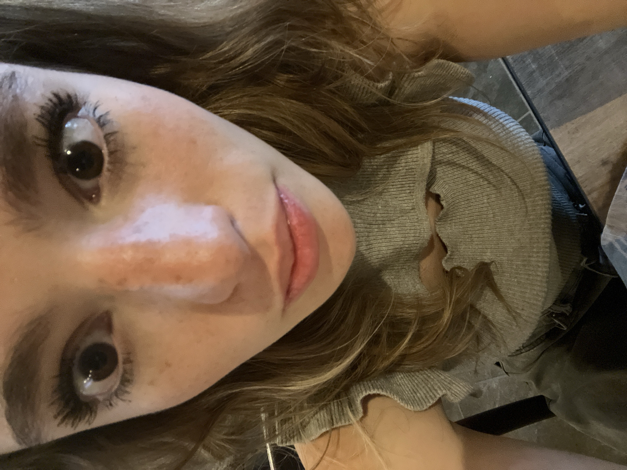 A close-up, angled photograph shows a young woman with brown hair and fair skin. She has dark brown eyes with thick, dark eyelashes, and freckles are visible on her face. She is wearing a gray, ribbed sweater with a ruffled collar. The background is slightly out of focus, showing a wooden surface. The photograph was taken indoors, but there is no information in the image to determine the location as San Bernardino Tlaxcalancingo, Mexico.