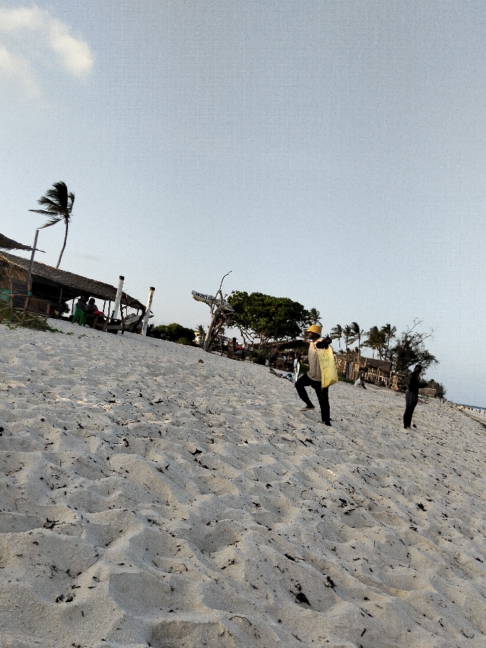 The scene depicts a sandy beach or dune environment in Gombato Bongwe ward, Kenya, viewed from a low angle looking uphill. The foreground and mid-ground are dominated by light-colored sand, showing numerous footprints and scattered dark organic matter.
In the mid-ground, a person wearing a yellow hat, a light-colored long-sleeved shirt, dark pants, and dark footwear is walking uphill from the right side of the frame towards the left. A light yellow or beige bag is carried on their back. To their right, further uphill, another individual is partially visible, dressed in dark clothing.
In the background on the left, a structure with a distinct thatched roof is visible, supported by wooden poles. Underneath this open-air structure, several individuals are seated. Adjacent to the structure, a tall palm tree with fronds bending left, indicative of wind, stands against the sky. Further right in the background, a dense cluster of green foliage and additional palm trees are present. A light-colored wooden or driftwood-like structure with illegible text (partially visible as "WAT") is positioned near the foliage.
The sky is a light, clear blue, with a single small white cloud visible in the upper left quadrant. The overall lighting suggests either early morning or late afternoon.