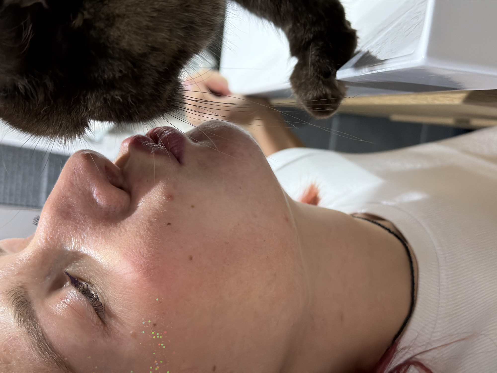 A photograph taken indoors depicts a woman lying on her back, with a dark-furred animal, possibly a cat, positioned above her head. The animal's paw is visible near the woman's face. The woman has fair skin with blemishes and glitter around her eye. She is wearing a white top. The background is blurred and consists of a white surface and a gray wall. There is no information present in the image to indicate the location as the city of Chaikovsky, Russia.