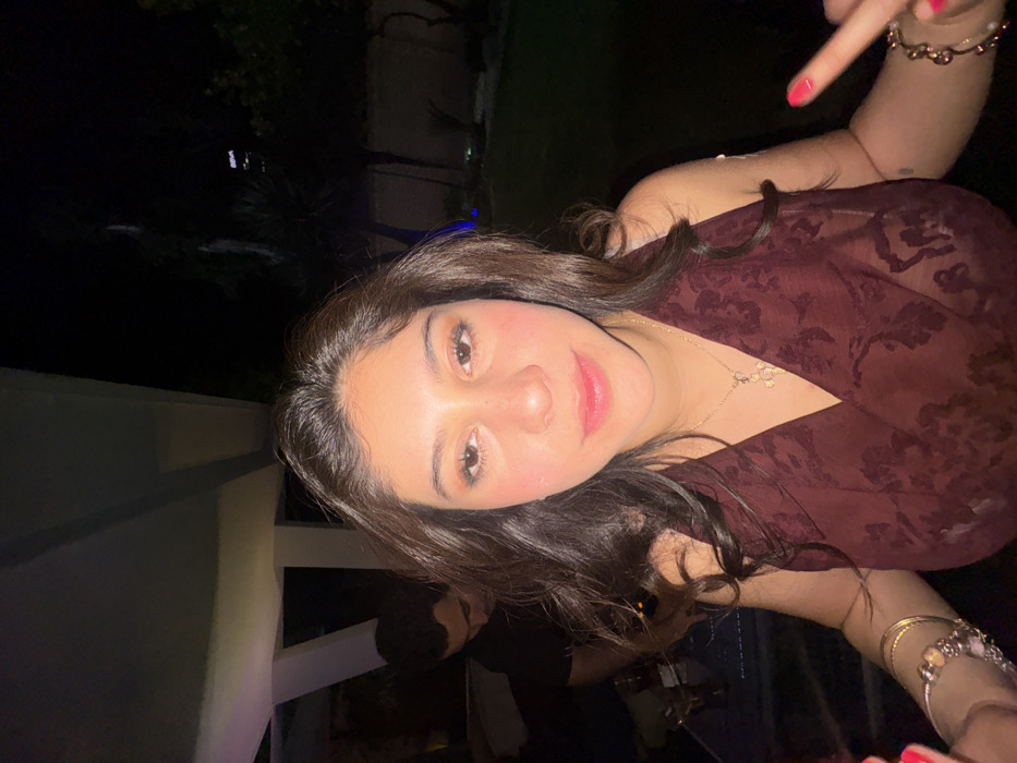 The image displays a nighttime scene in San Bernardino Tlaxcalancingo, Mexico, featuring two individuals in an outdoor or semi-enclosed patio setting.
The primary subject, a young adult female, occupies the foreground, looking directly at the camera. She has long dark brown hair and is wearing a dark red, patterned halter-neck top. A gold chain necklace with a light-colored pendant is visible around her neck, and multiple bracelets adorn her left wrist. Her fingernails are painted bright pink. Her left hand is raised, with the middle finger extended.
In the blurred left background, a young adult male is visible from the chest up, looking downward and away from the camera. He wears a dark shirt and is positioned near a dark surface, likely a table, on which bottles and glasses are discernible.
The environment includes a light-colored overhead structure or patio roof supported by white columns visible in the upper left. Beyond the subjects, a dark garden area with green foliage is present, with a faint blue light source illuminating a section of the ground. In the far distance, against the dark night sky, an illuminated building displaying the "BBVA" logo is visible. The scene is illuminated by artificial light, indicative of nighttime photography with a flash.