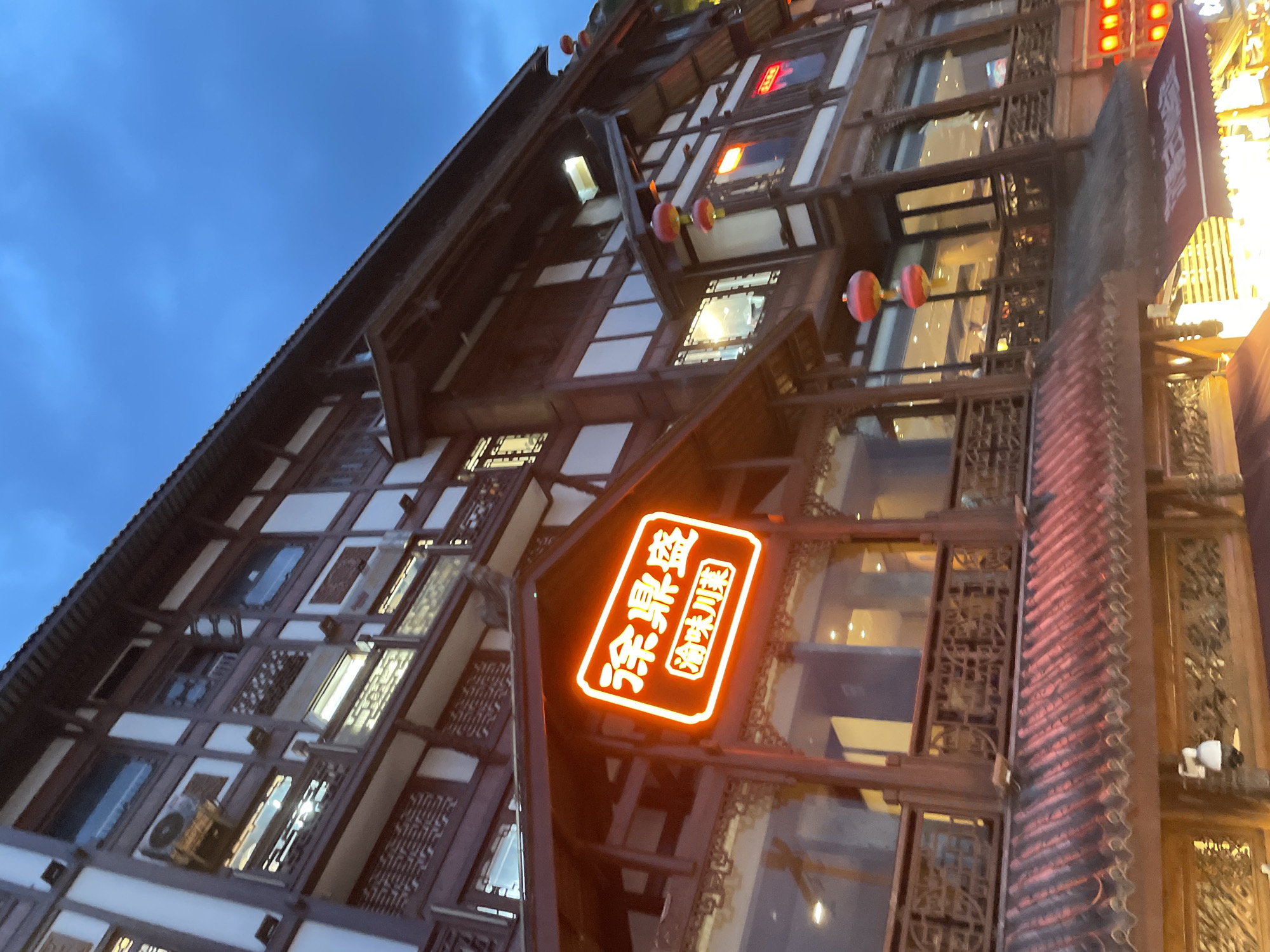 An exterior, low-angle, medium shot captures a traditional Chinese building with a neon sign in Yuzhong District, China. The building features dark wood accents and white paneling, with multiple stories and large windows. Red lanterns are visible hanging from the building. The neon sign, a bright orange rectangle, is positioned prominently on the building's facade and displays Chinese characters. The sky above is a deep blue, suggesting it is either dusk or night. The overall impression is one of an illuminated establishment in an urban setting, likely a restaurant or shop, designed in a traditional architectural style.