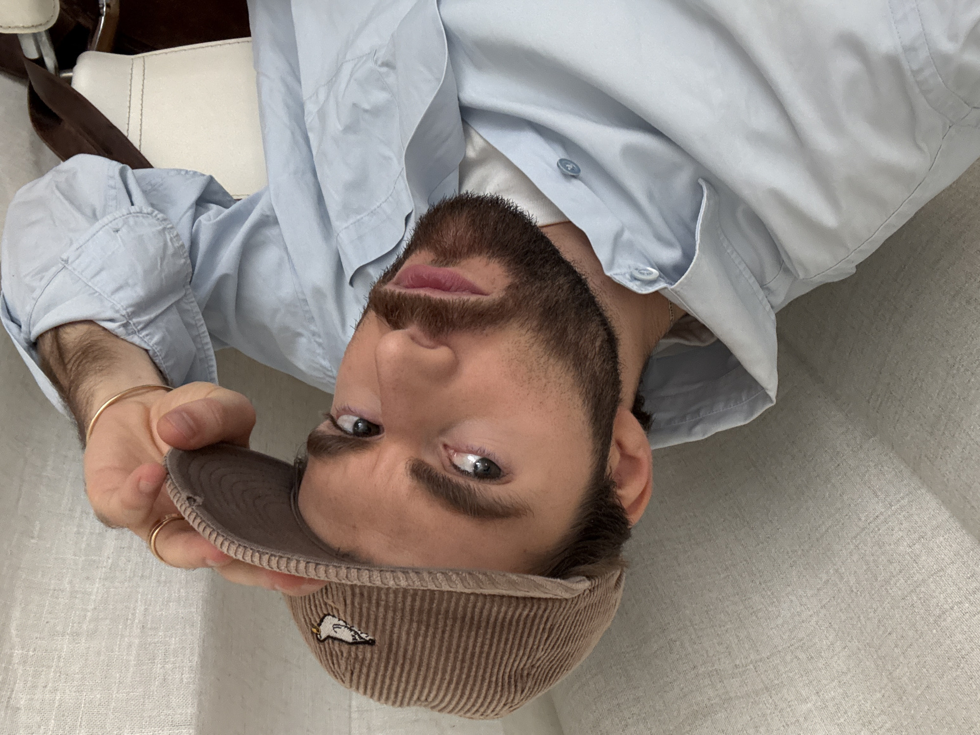 A man is photographed in an upside-down orientation, wearing a light blue collared shirt and a brown corduroy cap. He has a beard and is wearing a gold ring and a gold bangle on his left hand. The background consists of a white surface, possibly a couch or chair, and a beige textured wall. The image appears to be taken indoors, with natural light illuminating the scene. There is no indication of this image being from Montreal, Canada.