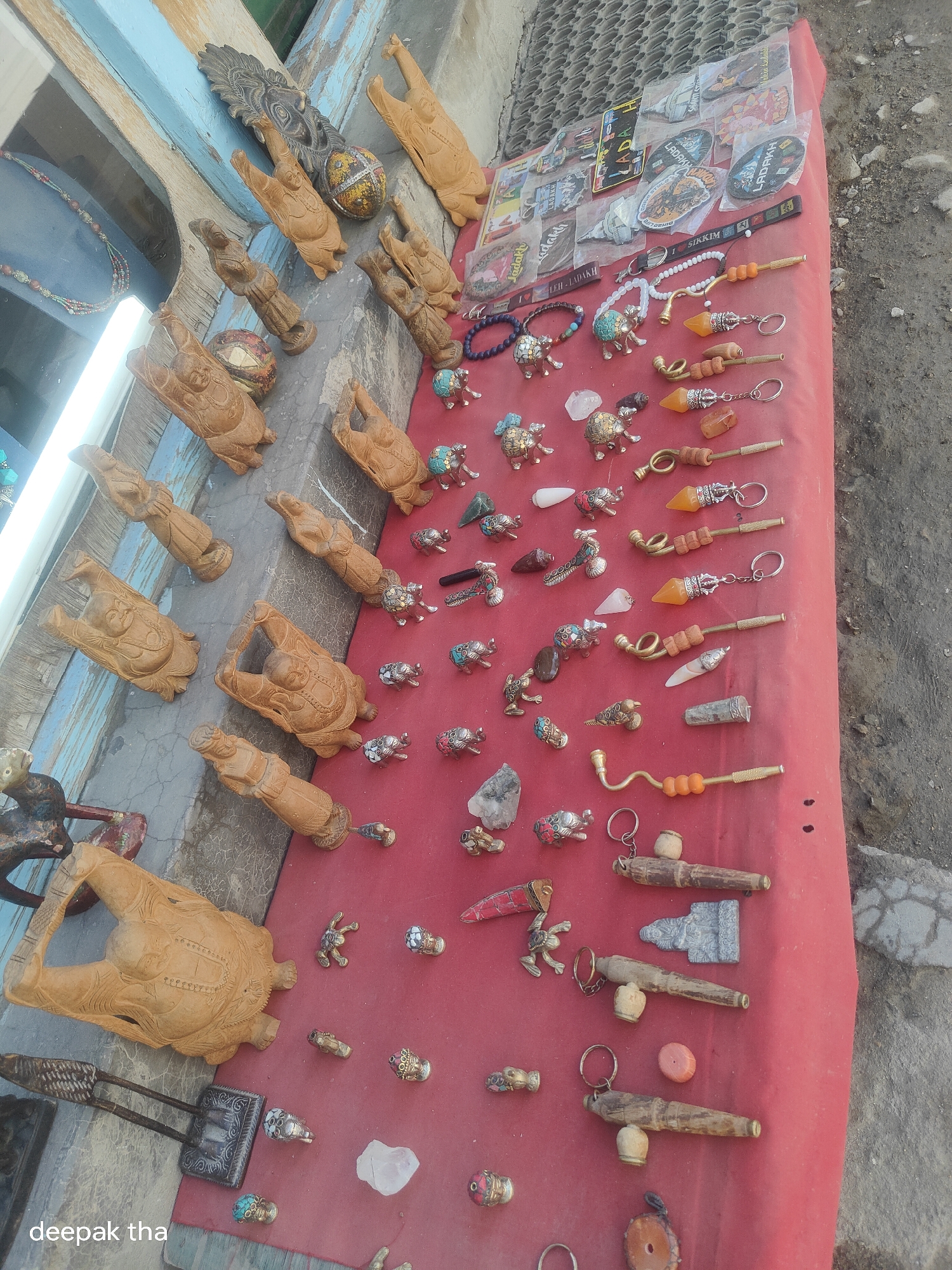 This is an overhead, high-angle, eye-level shot of a market stall in Leh, India, displaying various handicrafts for sale. The stall is covered with a red cloth and features a variety of carved wooden figures, keychains, and other trinkets. The wooden carvings appear to be of animals like elephants and turtles, as well as human figures. There are also several keychains with colorful beads and small decorative elements. In the background, a textured grey surface suggests a paved area, possibly a street or pathway. The lighting is natural, casting soft shadows, and the overall scene suggests a bustling outdoor market environment. The stall is positioned outdoors, with a glimpse of a building structure with blue painted wood on the left side. A watermark "deepak tha" is visible in the bottom left corner of the image.