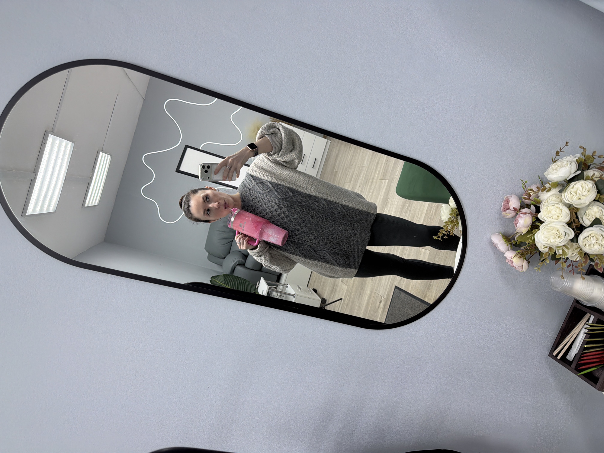 A woman is taking a mirror selfie, holding a pink cup and a white iPhone. She is wearing a gray sweater and black leggings. The mirror is oval-shaped with a black frame, reflecting an interior with a gray sofa and white cabinets. The wall next to the mirror is light gray and decorated with a bouquet of white and pink flowers, disposable cups, and a wooden box with stirring sticks and napkins. There are two rectangular LED lights visible in the reflection, suggesting an office environment. The photograph may have been taken in Perm, Russia, as part of a social media update.