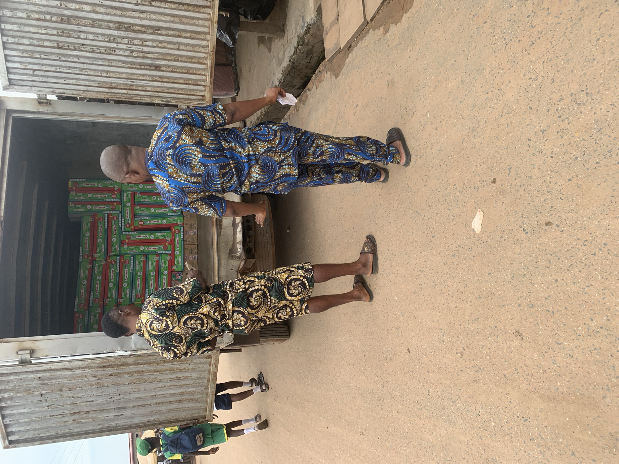 Two men, dressed in traditional Nigerian attire, are seen near the back of a truck loaded with boxes. The man in the foreground wears a green and gold patterned dashiki and shorts. He is facing away from the camera and appears to be looking into the truck. The second man, further back and to the left, is wearing a blue and gold patterned dashiki and trousers. He is also facing away from the camera, holding a small white object in his right hand.
The truck is open at the rear, revealing stacks of green and red boxes. The boxes have text on them, including "9 EXTRA" and what appears to be a logo. The ground is a dirt or asphalt surface, appearing somewhat dusty. In the background, parts of a building and a ditch are visible. The lighting suggests it is daytime. The overall scene depicts a commercial transaction or delivery, possibly involving the loading or unloading of goods. The people's postures do not convey specific emotions, but the presence of a truck and stacked boxes implies activity related to trade or logistics. The context provided places the scene in Shagamu, Nigeria.