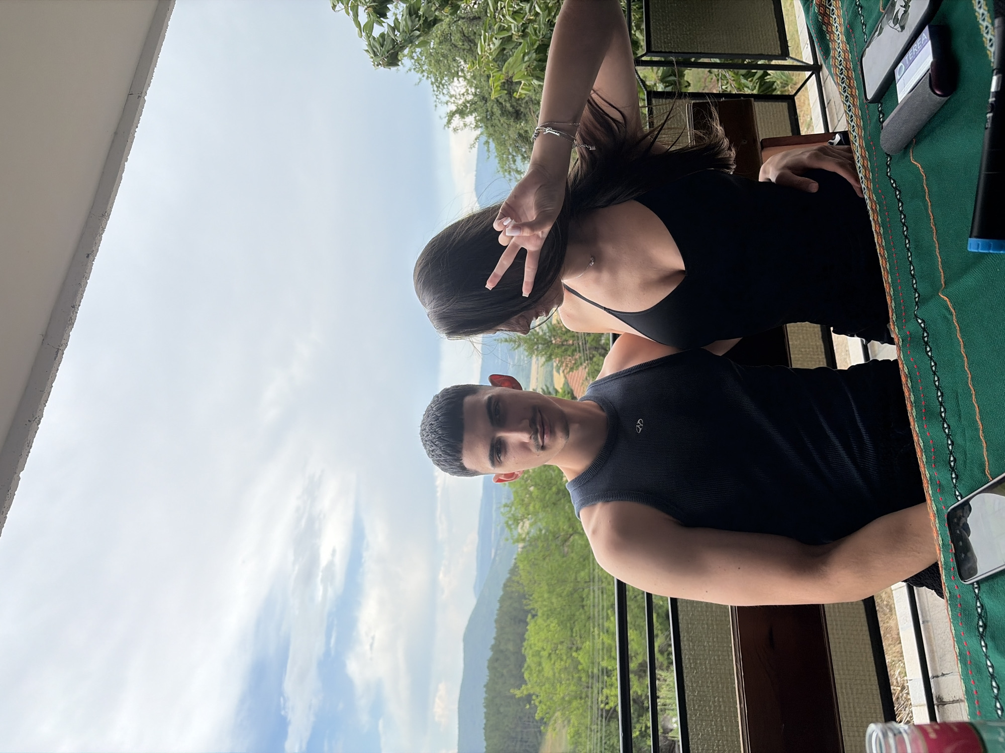 The media file shows two young adults, a woman and a man, on a balcony overlooking a lush, mountainous landscape. The woman, with long dark hair, is leaning over and making a peace sign with her right hand, her face obscured. She is wearing a black tank top and a silver bracelet. The man, in the foreground, is looking directly at the camera with a neutral expression. He has short, dark hair and a mustache, and is wearing a dark tank top. Behind them, the landscape is green with trees and rolling hills, suggesting a rural or mountainous location. The sky is partly cloudy, indicating daytime. On the table to the right, a green cloth with an embroidered pattern is visible, along with a smartphone displaying the text "TEREA". The overall scene suggests a casual outing or vacation. The presence of "TEREA" on the phone, combined with the location context of Vladislavtsi, Bulgaria, may indicate a connection to a specific product or service available in that region.