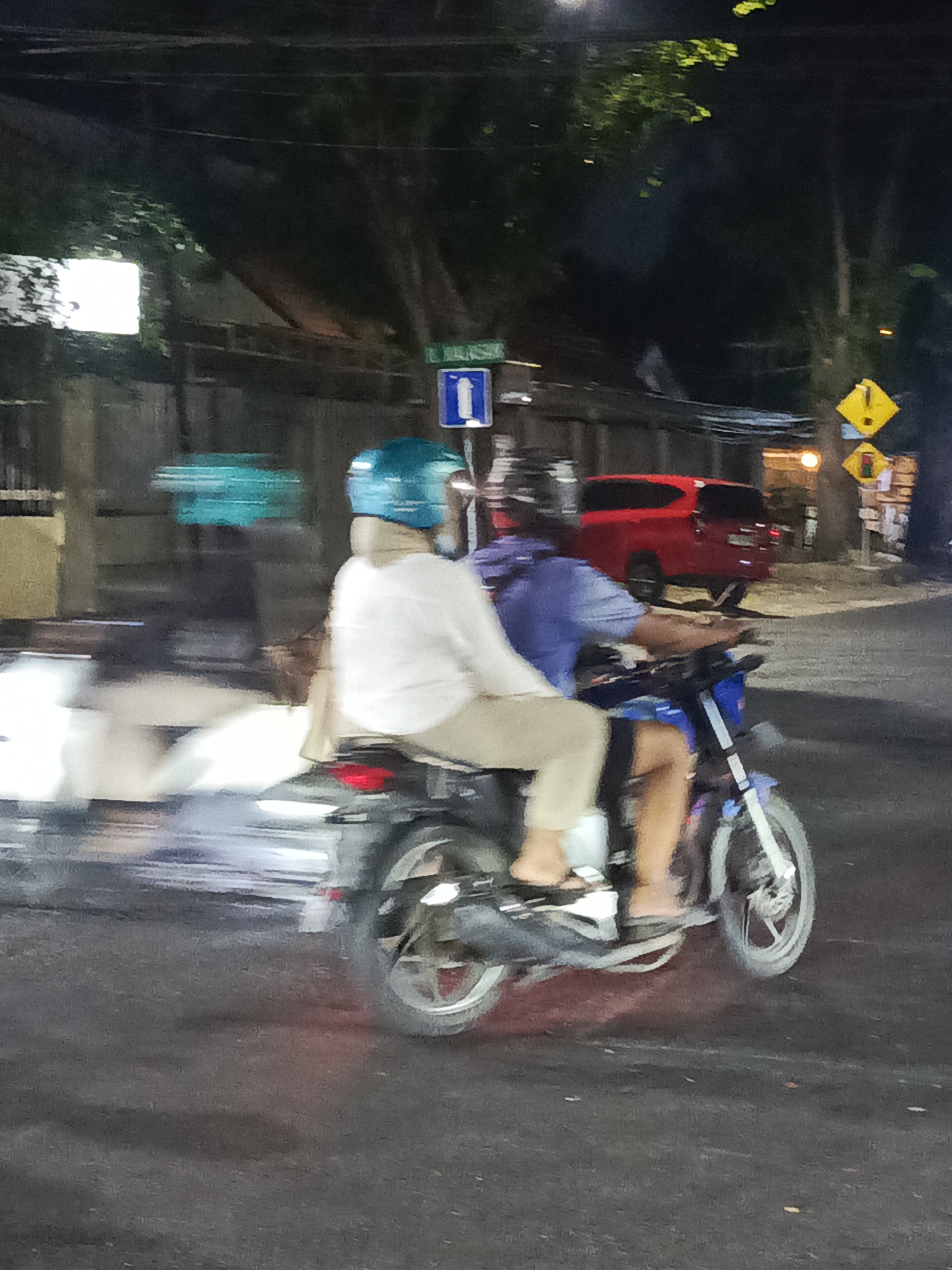 The image captures a nighttime street scene in Surabaya, Indonesia, with a focus on a motorcycle carrying two individuals. The motorcycle appears to be in motion, indicated by the motion blur of the background elements and the wheels. The rider in the front is wearing a turquoise helmet and a light-colored long-sleeved shirt and tan pants. The passenger behind them is wearing a blue shirt.
The environment includes a paved road, possibly wet due to the reflections and the blurred streaks of light. In the background, a red vehicle is visible, and there are street signs and lighting. Trees with foliage are also present, suggesting an urban or suburban setting. The overall impression is a snapshot of everyday life on the streets of Surabaya after dark.
