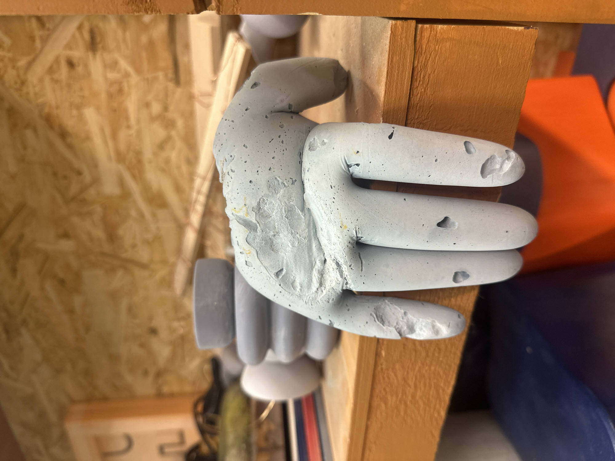 A close-up shows a gray, concrete hand-shaped object gripping a wooden plank. The hand has visible imperfections and damage, including chips and abrasions. The fingers are slightly curved and positioned as if grasping the plank tightly. The wooden plank is light brown and appears to be part of a larger structure or shelf. The background includes blurred elements of what looks like a workshop or storage area, with plywood walls and various objects out of focus. No information can be extracted to indicate the location is Moscow, Russia.