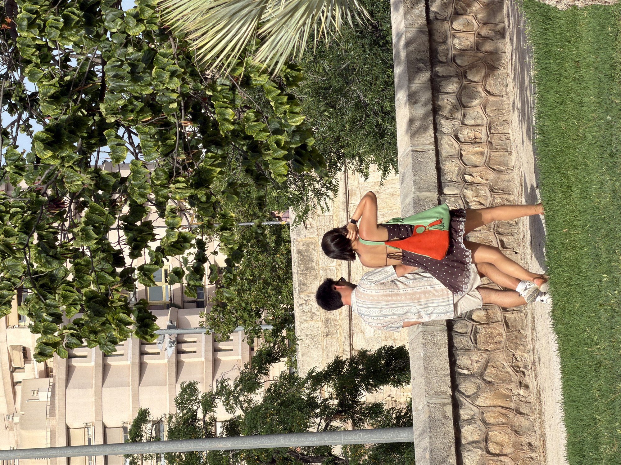 In an outdoor urban setting, a man and a woman are walking away from the camera, seen from behind and slightly to the side. They are positioned on a paved path behind a low, textured stone wall. The man is on the left, wearing a light-colored, patterned short-sleeved shirt and light-colored shorts. The woman is on the right, wearing a dark, patterned short dress with white trim and carrying two bags, one bright orange and the other green. She has her right arm raised and is touching the back of her head. The man is wearing white sneakers and the woman appears to be wearing sandals. To the left of the couple, a grey metal pole stands vertically. Behind the stone wall and the pathway, there is abundant green foliage from trees and shrubs. In the background, a large, multi-story building with a beige facade and visible windows is visible. The sky above is a clear blue. The scene is lit by daylight, casting shadows on the ground.