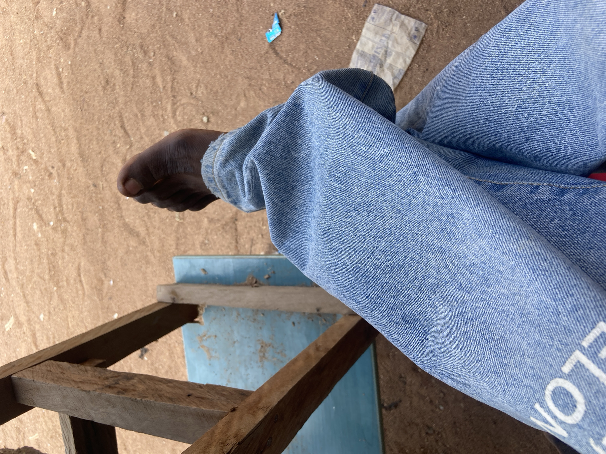 A close-up, horizontal, outdoor shot captures the lower body of a person wearing light blue denim jeans. The person's right leg is bent at the knee, with the foot resting on a rough, wooden structure. The foot is bare and dark-skinned. The background consists of sandy ground, with a light blue surface visible beneath the wooden structure, possibly a table or chair. A torn piece of white paper with blue markings is present on the ground behind the person's leg, along with a small, bright blue fragment. The sunlight suggests it is daytime. The jeans have white lettering that is partially visible and appears to spell "LLON". The setting appears to be informal and possibly rural, consistent with a location context of Kuson, Nigeria. The image focuses on the textures of the denim, wood, and sand, and the stark contrast between the person's bare foot and their jeans.