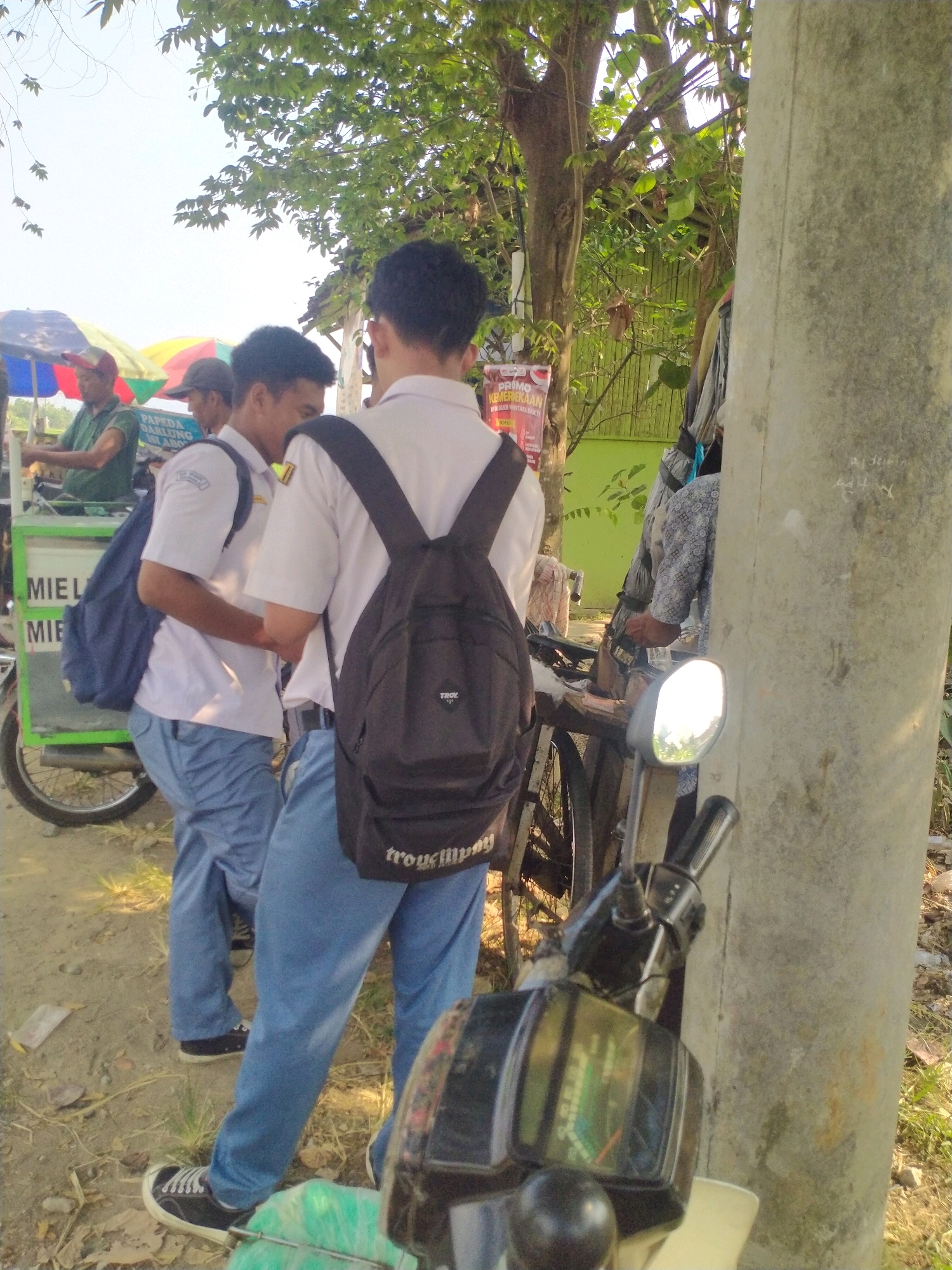 In Begajah, Indonesia, on a dirt road, two young males in white school uniforms and blue pants stand near a food cart and a motorcycle. One male, facing away, wears a black backpack and a white short-sleeved shirt. The other, also facing away, wears a navy blue backpack over a white short-sleeved shirt. A third male, wearing a green shirt and a red and yellow cap, is visible behind the food cart. The food cart has a green body and displays text in Indonesian. A motorcycle with a chrome mirror is parked in the foreground, and a concrete pole stands to the right of the scene. Trees and foliage create a backdrop.