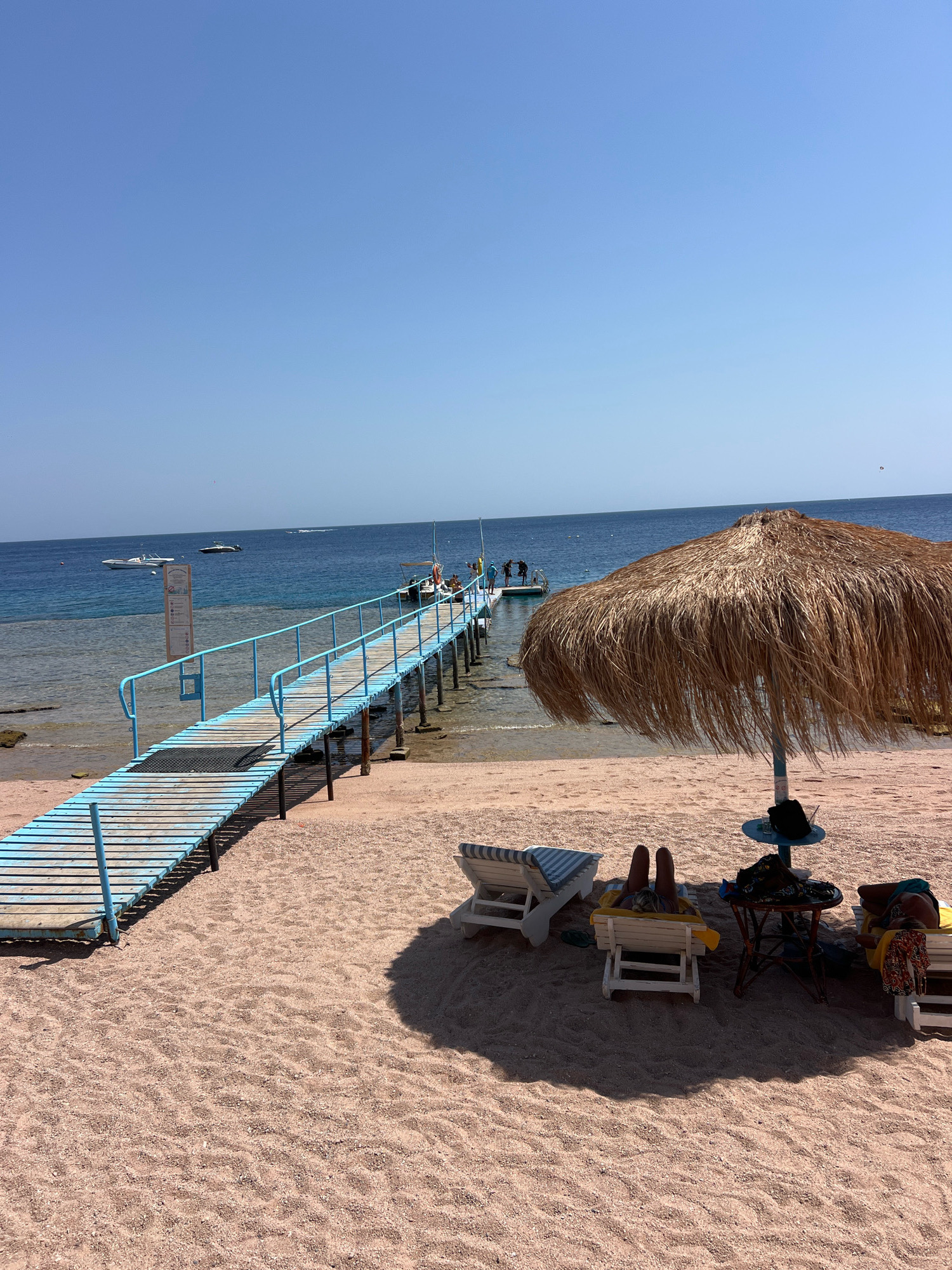 A bright, sunny day at a beach resort in Sharm El Sheikh, Egypt. A long wooden pier with light blue railings extends into the clear blue sea. Several boats are visible in the water, with some people standing on the pier. On the sandy beach, there are lounge chairs, one with two people relaxing, and a straw umbrella providing shade. The sand is a pale pinkish-brown color. The sky is a clear, uniform light blue.