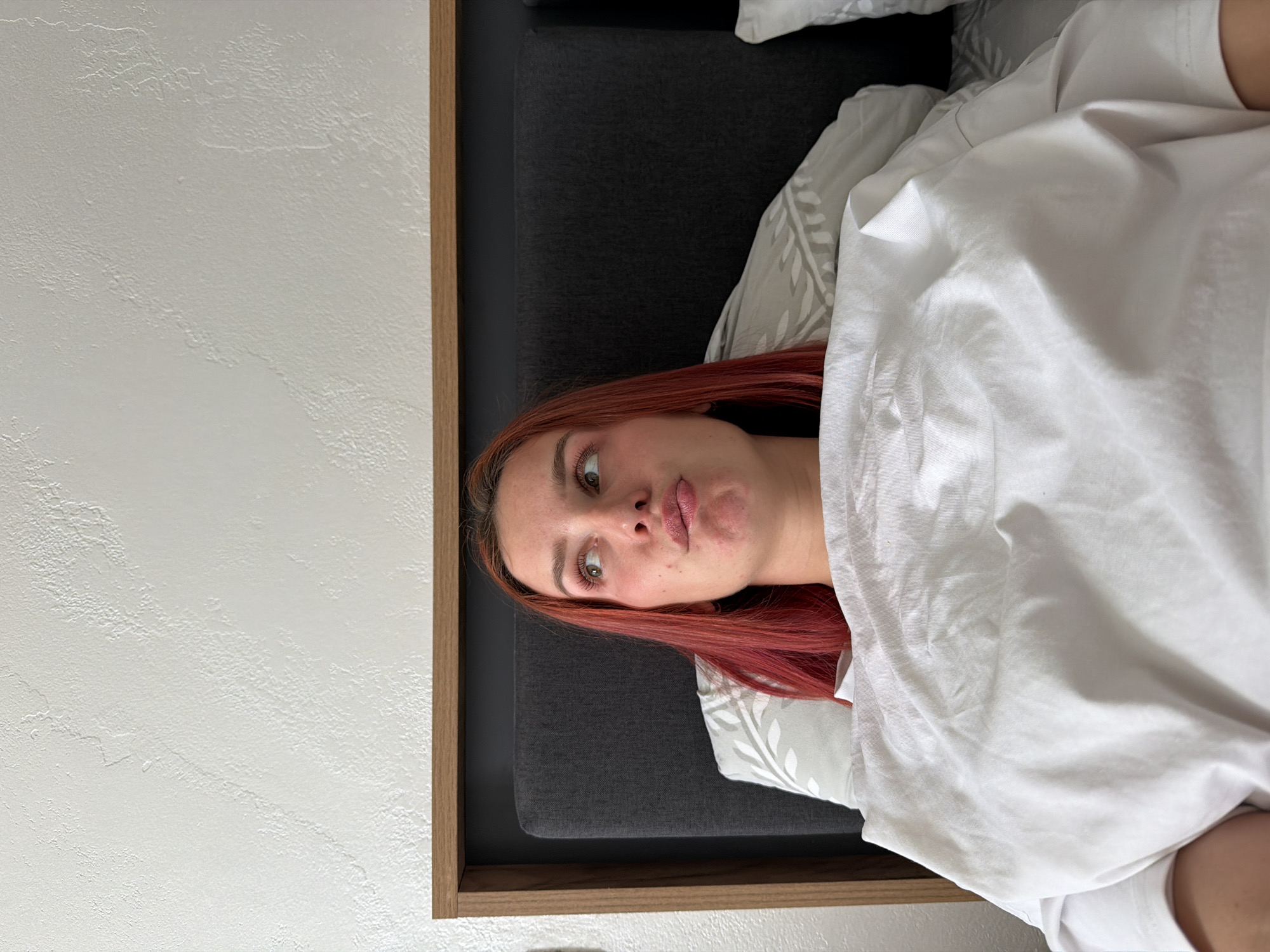 A person with reddish hair is lying in bed under a white blanket. The person's face is visible, and they are making a slight pout. The bed has a gray headboard and pillows with a leaf pattern. To the left of the bed is a white wall with a textured surface. The image appears to be taken indoors. There is no information to suggest the photo was taken in Naberezhnye Chelny, Russia.