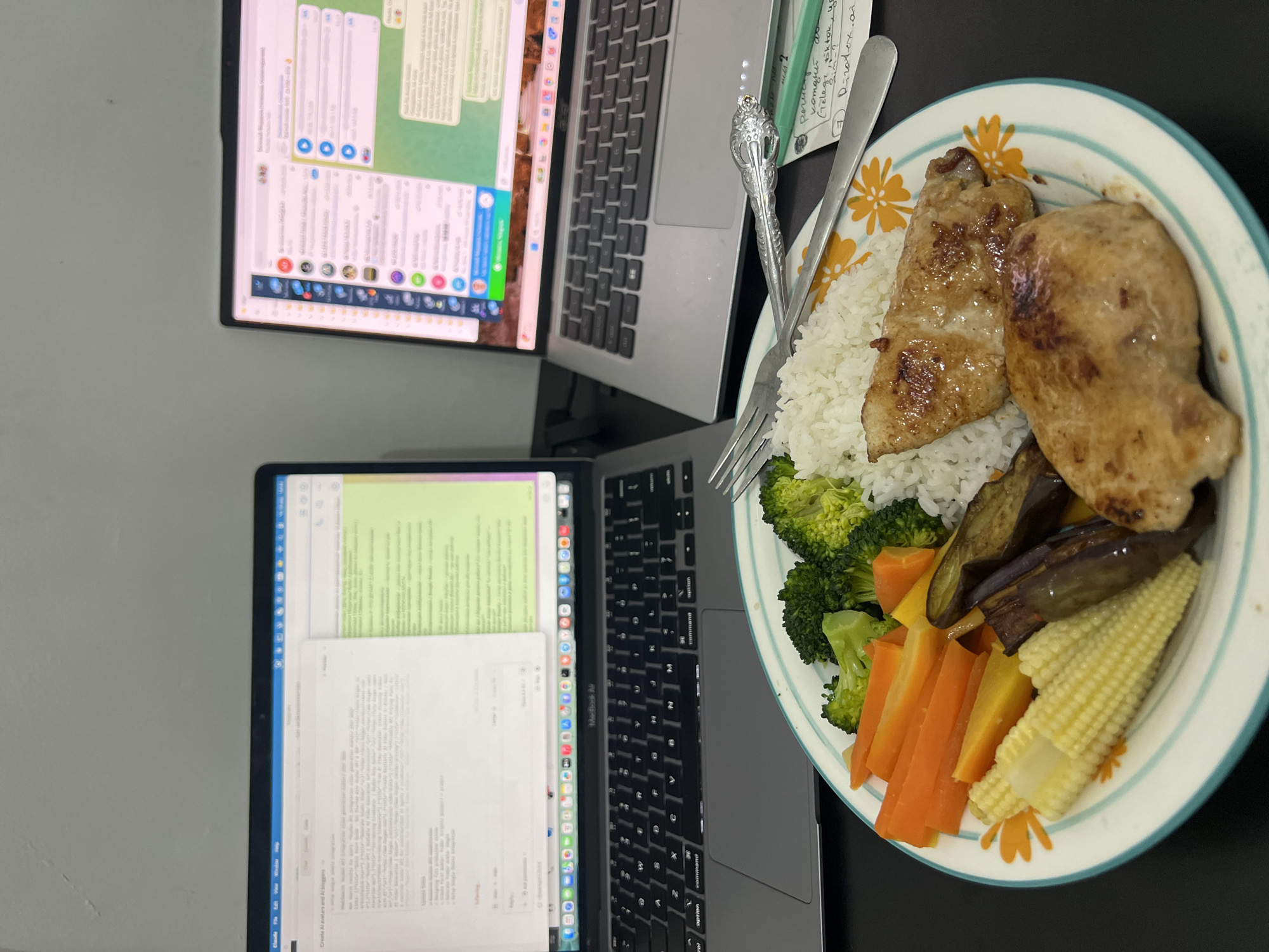 The image shows a desk with two laptops and a plate of food. The food on the plate consists of rice, two pieces of chicken, broccoli, carrots, baby corn, and eggplant. There is a fork and spoon next to the plate. The location of Celuk, Indonesia is not supported by image. The picture does not offer enough information to definitively confirm either a subject or action in this image.