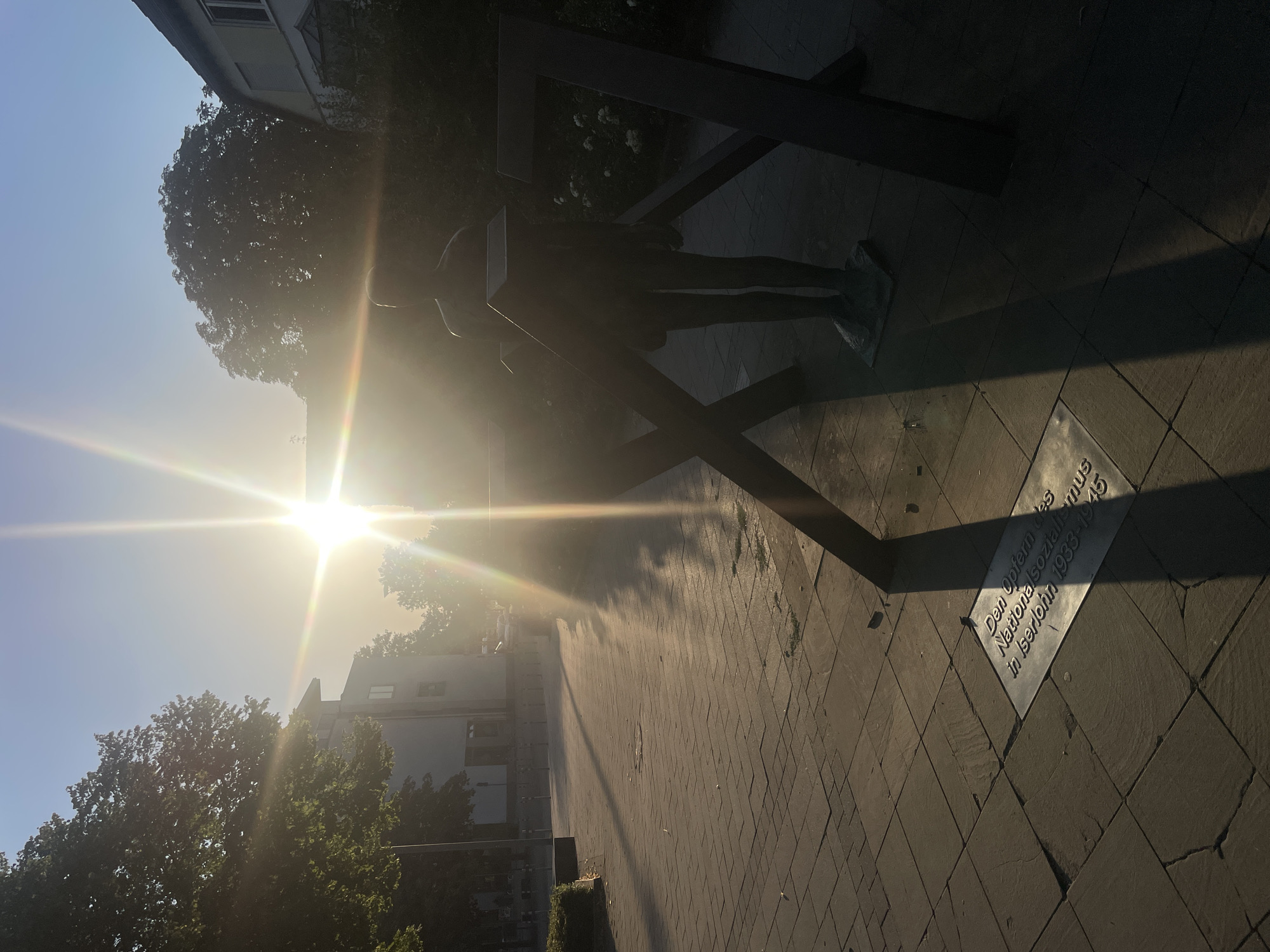 The photograph depicts a memorial located in Iserlohn, Germany, dedicated to the victims of National Socialism from 1933-1945. The sun, positioned high in the sky and slightly off-center to the left, casts a strong glare across the scene, creating lens flares and significant shadows. In the foreground, a bronze statue of a standing figure is partially obscured by abstract, dark metal structures resembling an "X" and a "7". A plaque inscribed with German text is visible on the paved ground in front of the statue. The surrounding environment includes buildings and trees silhouetted against the bright sky, indicating an outdoor urban setting. The pavement is made of large, rectangular stone slabs.