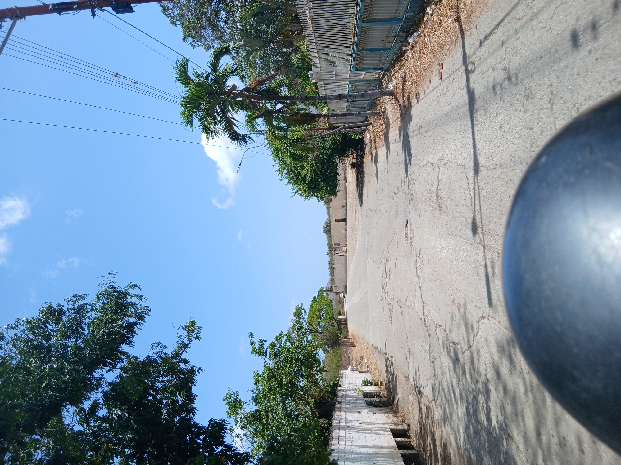 The image presents a street in Santa Ana, Venezuela, under a clear, blue sky with scattered white clouds. The road, made of cracked asphalt, stretches into the distance. On the right side of the road is a weathered fence made of white wood with blue trim, behind which lie trees with bare branches. On the left side of the street, there are trees with dense, green foliage and a wall made of white concrete. Utility poles carrying wires are visible against the sky. The overall lighting suggests a bright, sunny day. A portion of a gray circular object, possibly part of a vehicle, occupies the lower right corner of the frame.